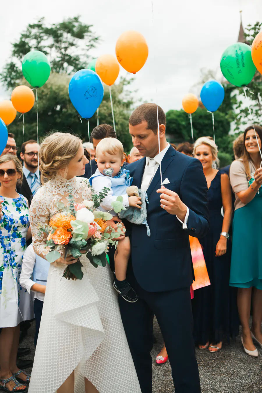 A groom holds a baby with a pacifier in front of a bride in a white dress, surrounded by people holding colorful balloons.