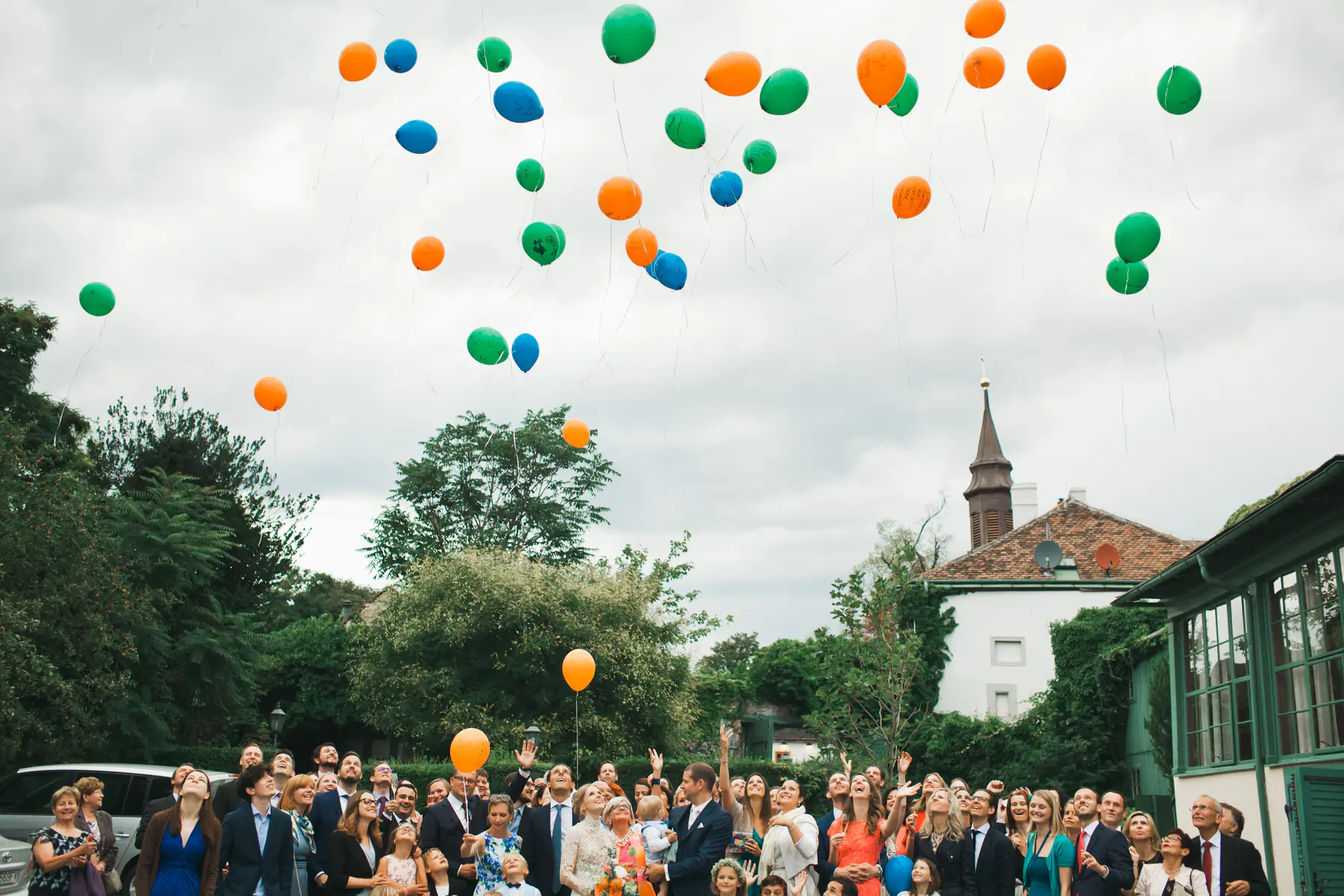A large group of wedding guests looks up at dozens of colorful orange, green, and blue balloons floating into an overcast sky, with a church steeple visible in the background.