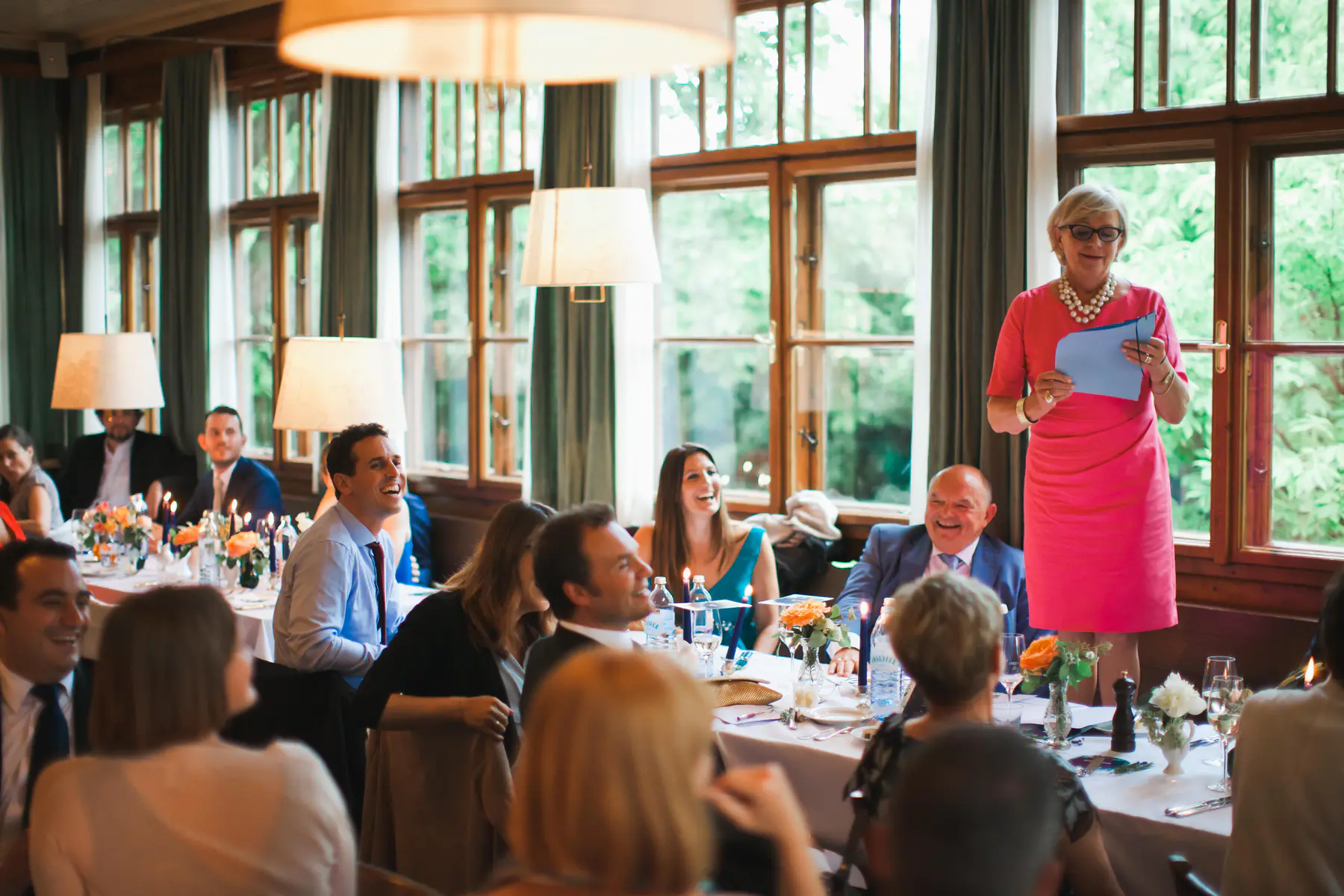 Candid wedding moments at historic Pfarrwirt restaurant. An older woman in a bright pink dress and pearl necklace stands smiling, holding a blue card, as she addresses a room full of people seated at tables. Many guests are smiling and laughing, looking towards her. The tables
