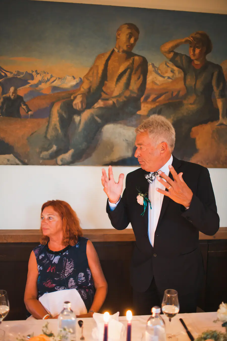 An older man in a black suit with a pink boutonniere and bow tie speaks while gesturing with both hands. An older woman with red hair sits at a table with lit candles next to him.
