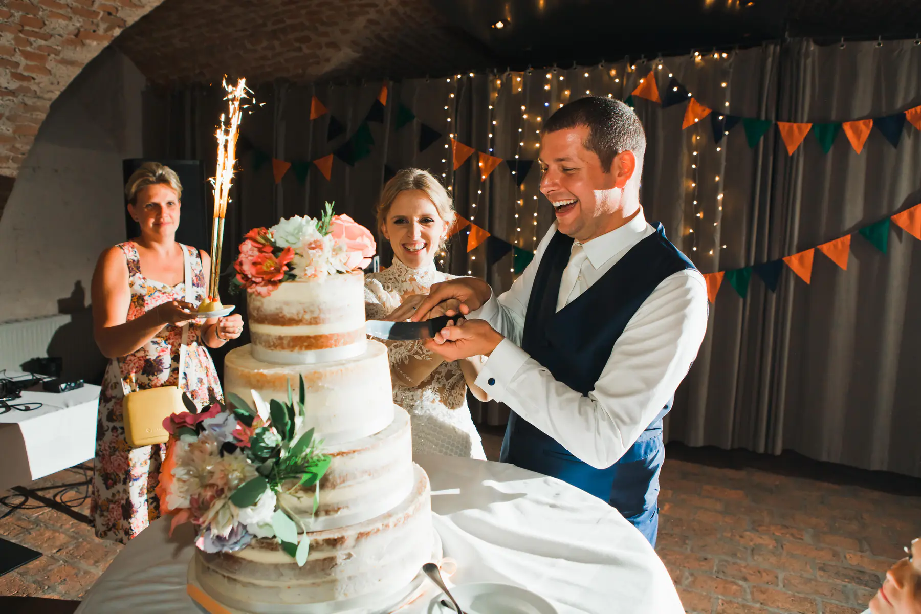 A smiling bride and groom cut a multi-tiered wedding cake decorated with flowers. A woman stands beside them, holding a sparkler. String lights and festive bunting are visible in the background.