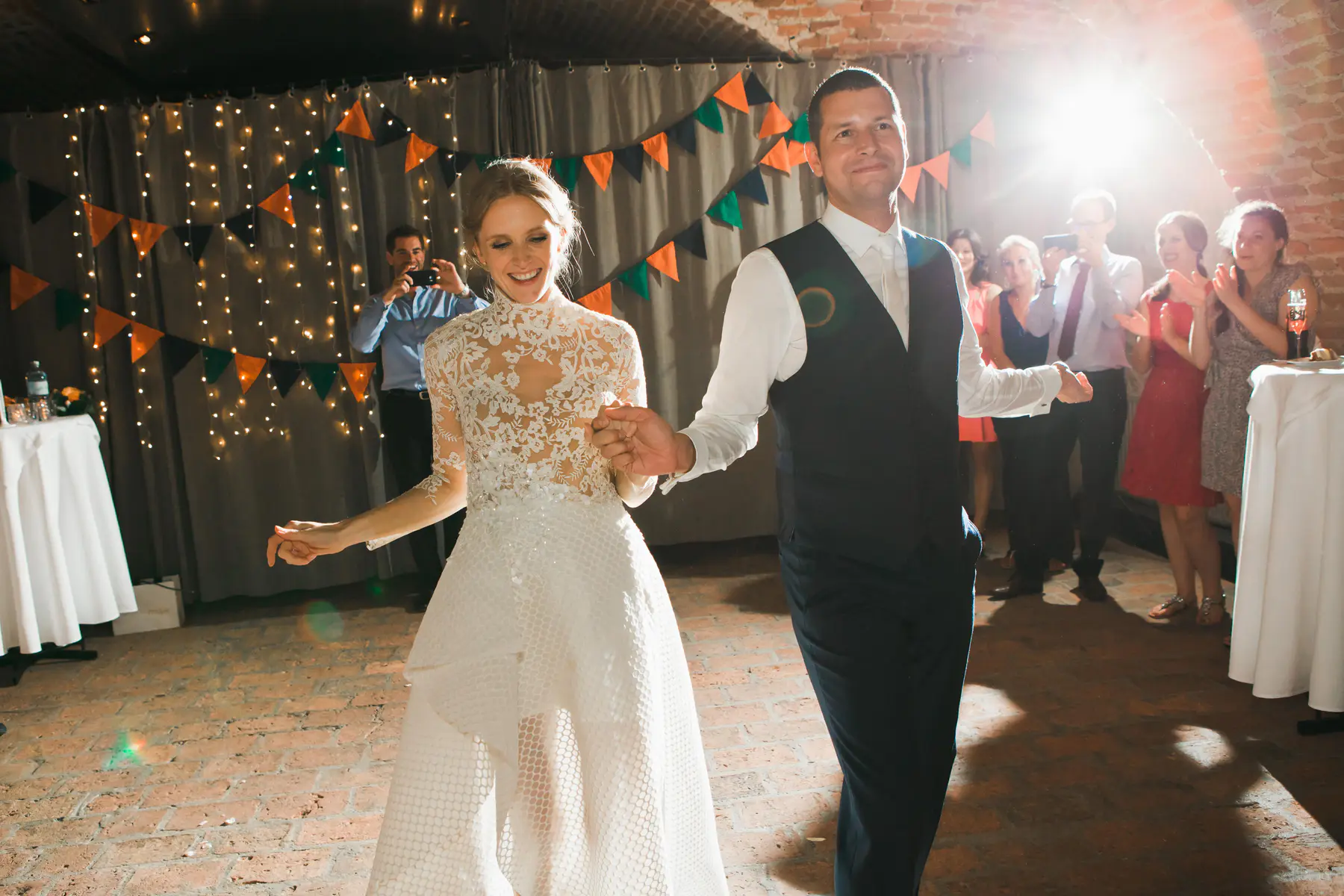 A smiling bride in a white lace dress and groom in a dark vest dance hand-in-hand, illuminated by a bright light. String lights, pennant banners, and clapping guests
