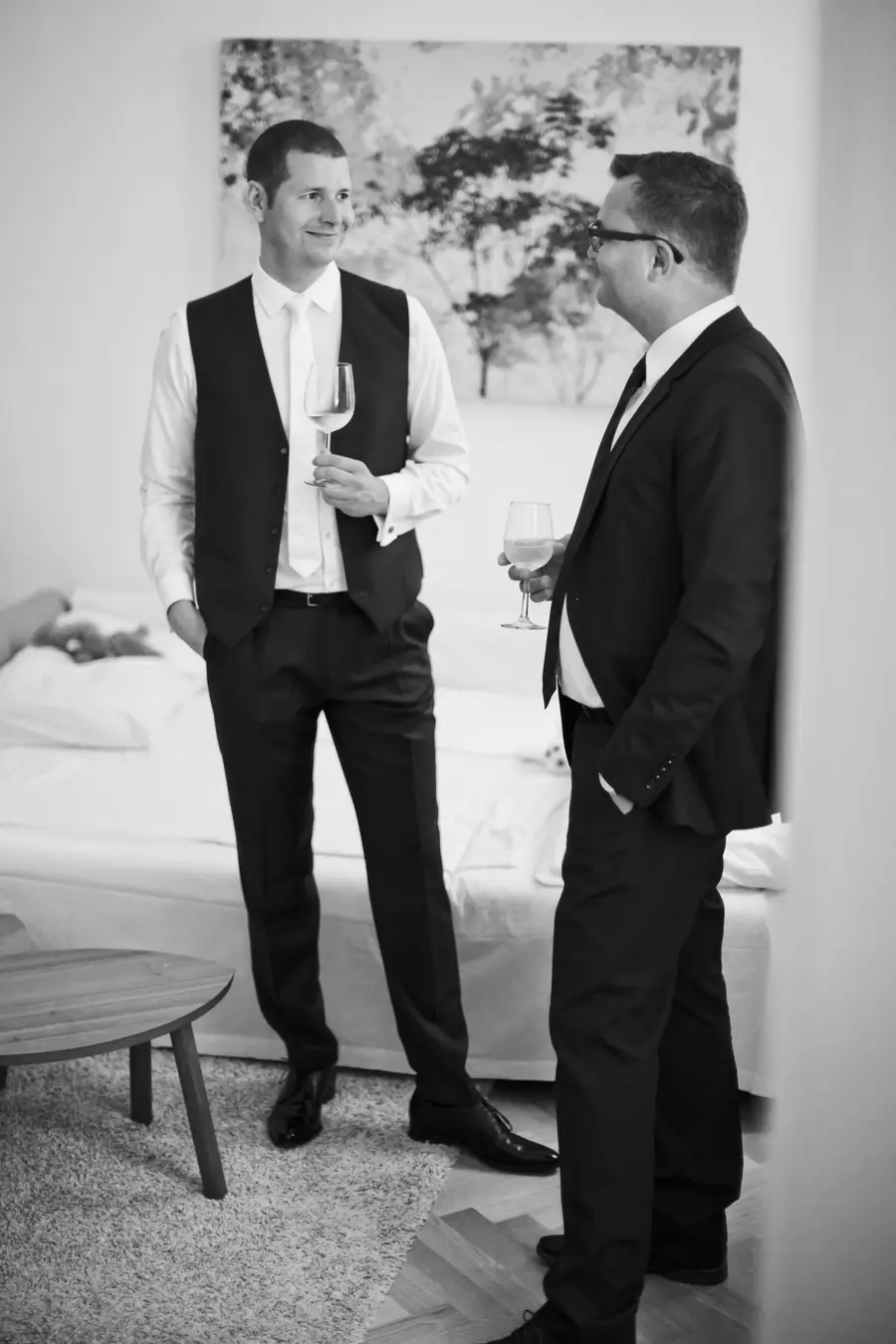 A man in a suit and vest with a glass of wine talks to another man in a suit holding a glass of wine.