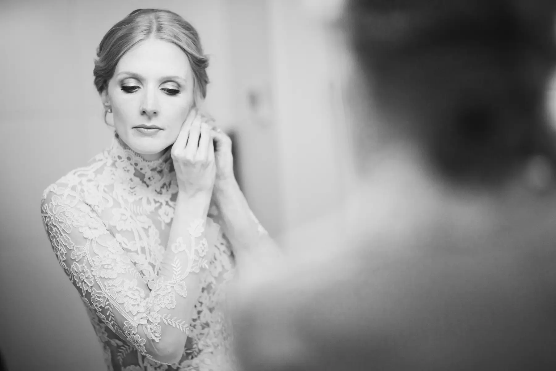 A bride in a lace dress adjusts her earrings while looking down, with her reflection visible in a mirror.