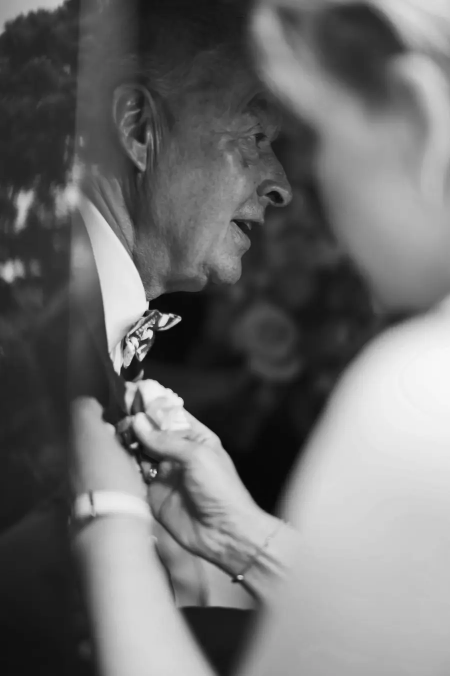A black and white close-up shows an older man in profile, wearing a white shirt and patterned bow tie, with his mouth slightly open. Another person's hands are in the foreground,