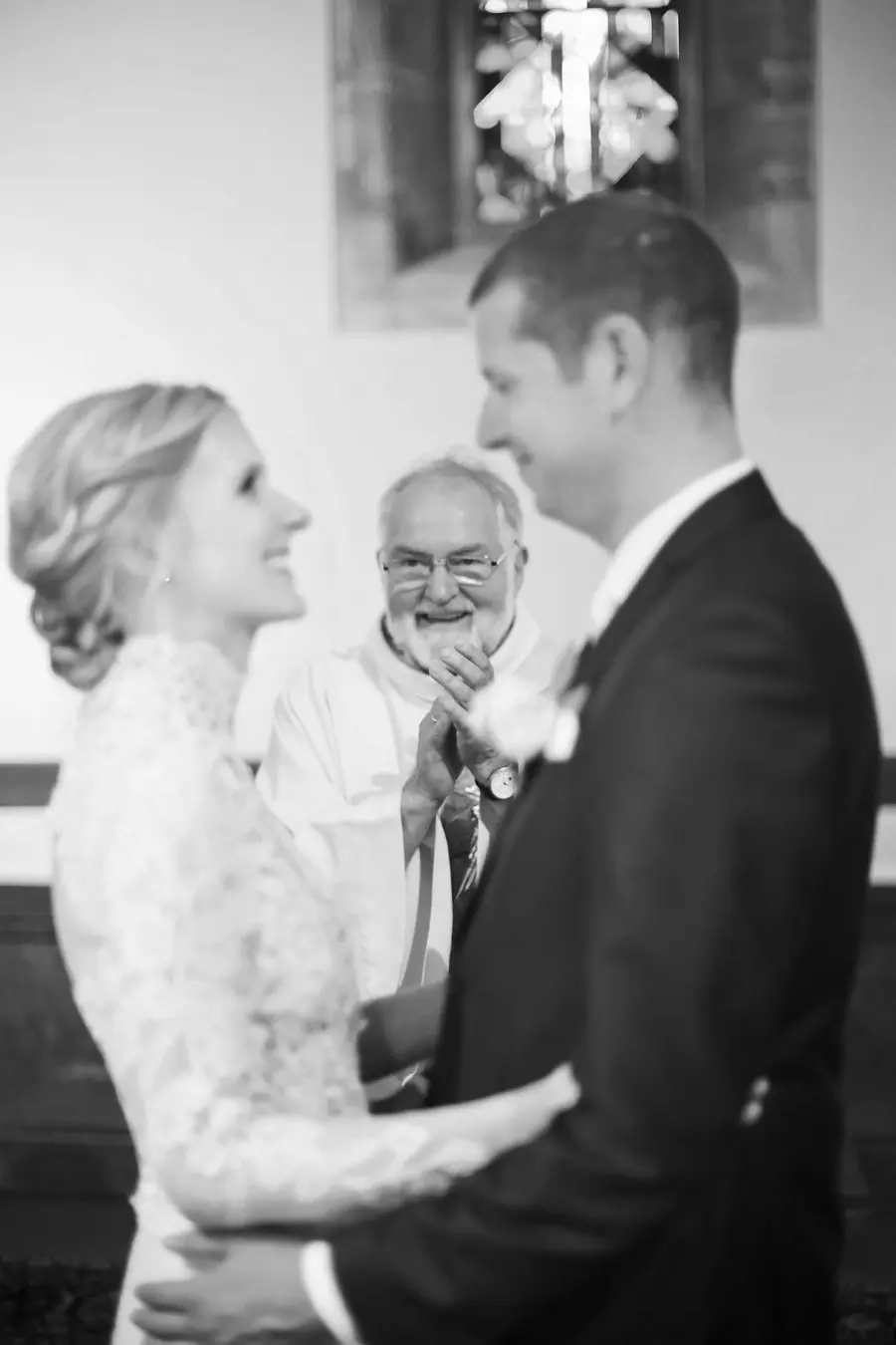 A brief, accurate description for alt text accessibility:

**Black and white photo of a smiling bride and groom looking at each other during their wedding ceremony, with a beaming, bearded officiant