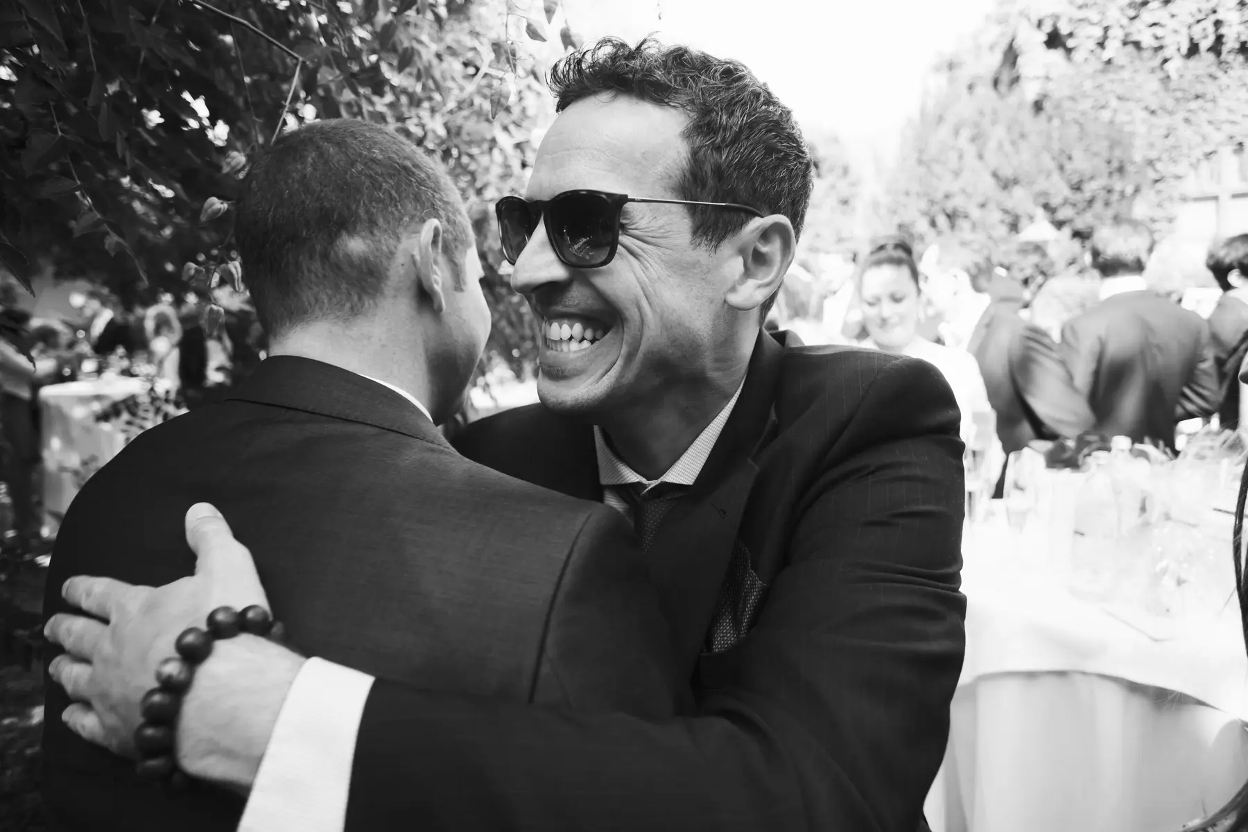 A black and white photo of two men embracing. The man on the right is wearing sunglasses and is laughing heartily with his mouth open wide. The man on the left is seen from behind, with his arm around the other man's shoulder and a beaded bracelet on his wrist.