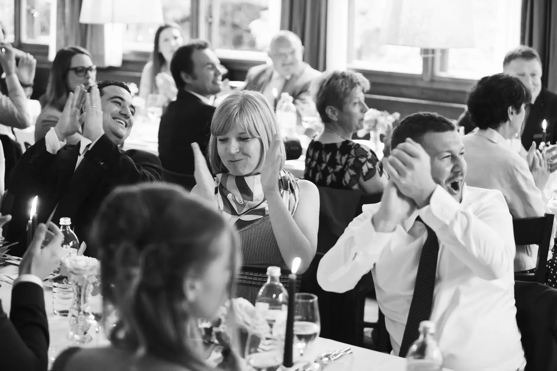 A black and white photograph shows a lively indoor gathering where many guests are seated around tables, enthusiastically clapping and smiling or laughing. Lit candles adorn the tables.