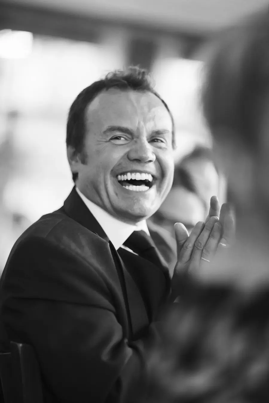 A black and white photo shows a man in a suit laughing heartily, with his mouth wide open, while clapping his hands.