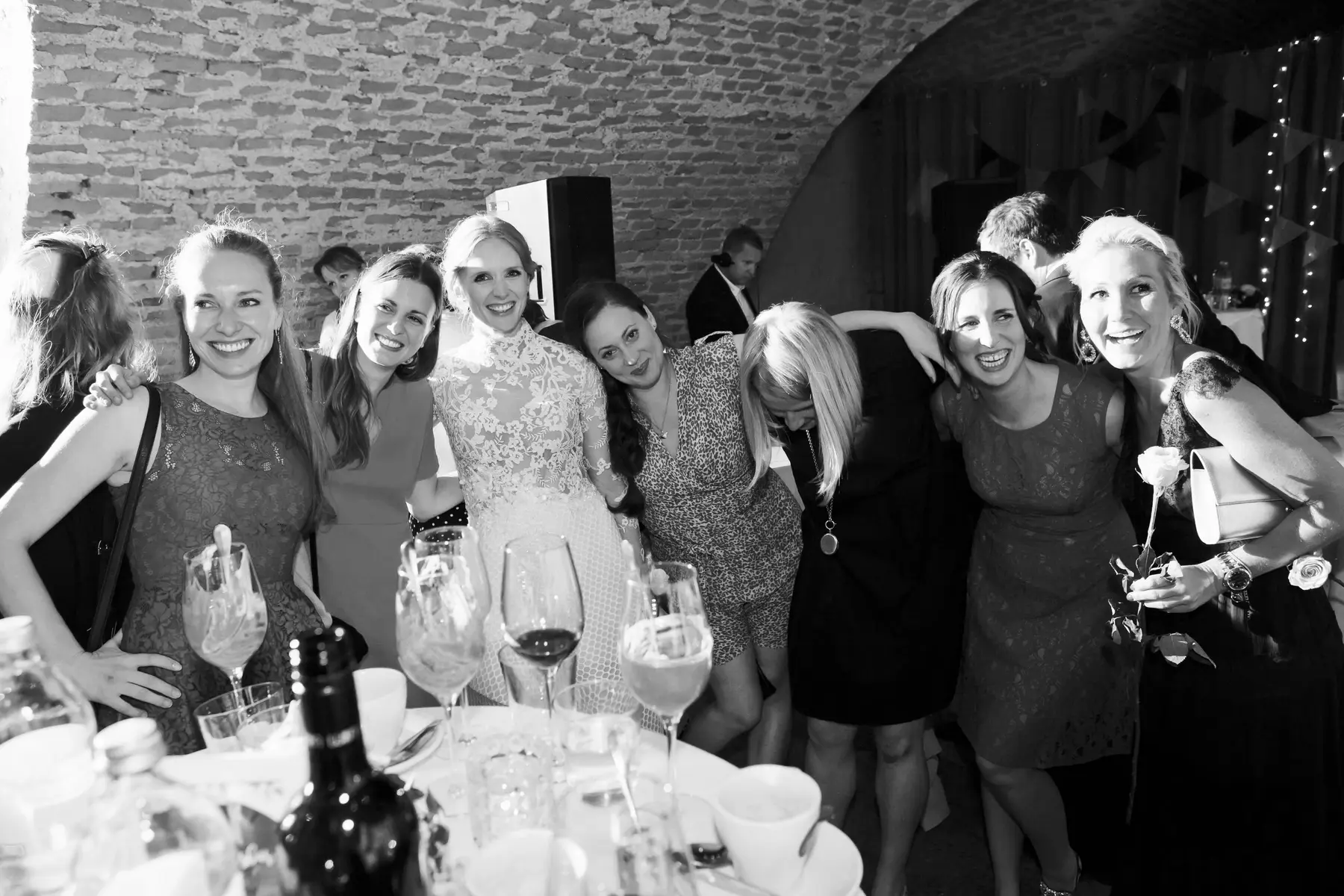 Candid wedding photography Vienna - natural reportage style. A black and white photo shows a joyful group of eight women posing together at a festive event. They are smiling and laughing around a table laden with drinks in a rustic venue with brick walls and