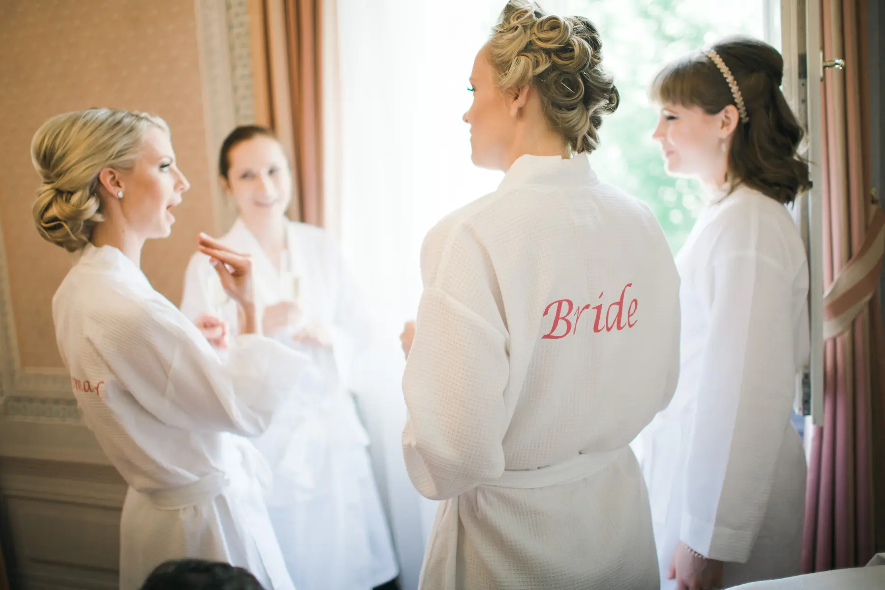 A bride with her hair in an elegant updo is being hugged by a woman in a white dress, with a third woman in the background attending to makeup.