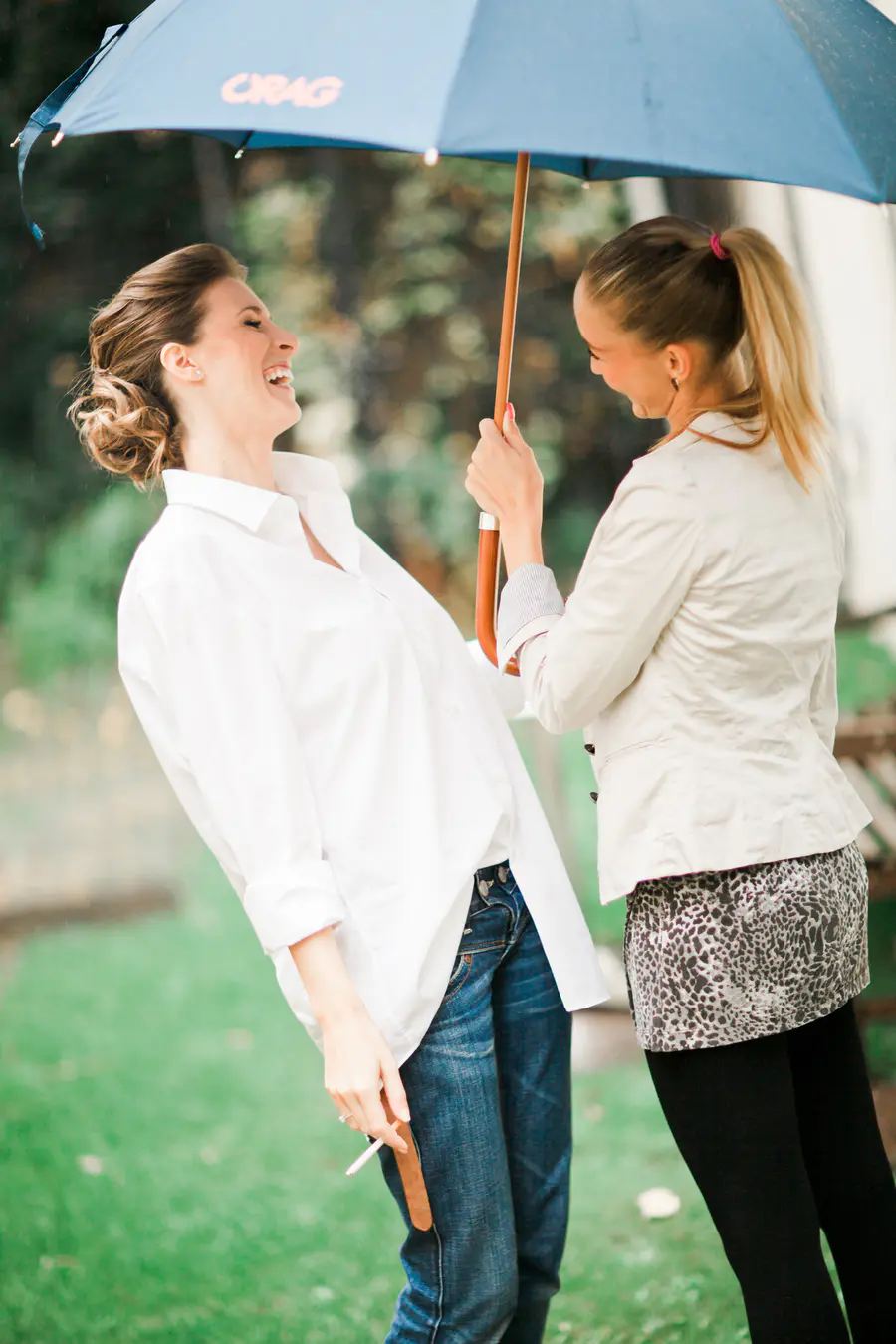 Two women share a moment of laughter outdoors under a blue umbrella. The woman on the left, with hair in a bun, leans back with a wide smile, wearing a white shirt and