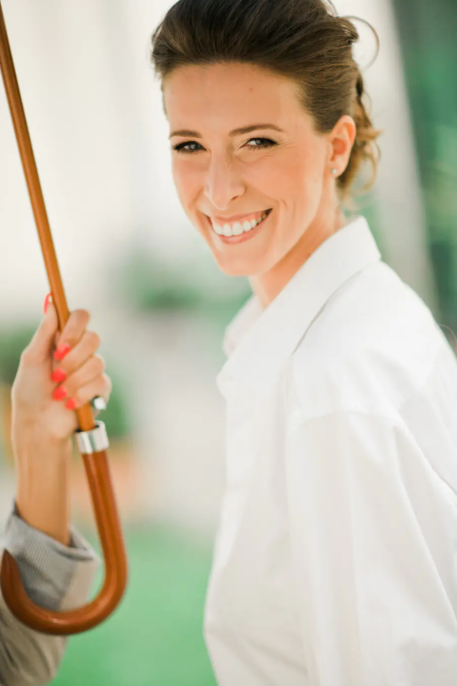 A smiling woman in a white shirt holds a curved wooden umbrella handle, with bright coral nail polish.