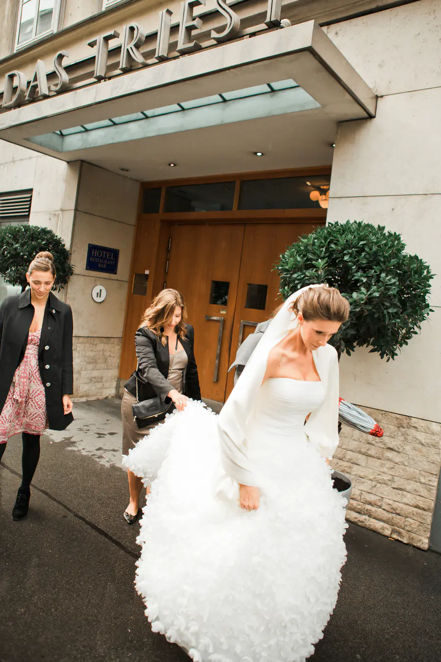 A bride in a voluminous white strapless wedding dress with textured, petal-like details is escorted from a hotel entrance by two women, one of whom is helping to carry her long skirt. At hotel Triest in Vienna