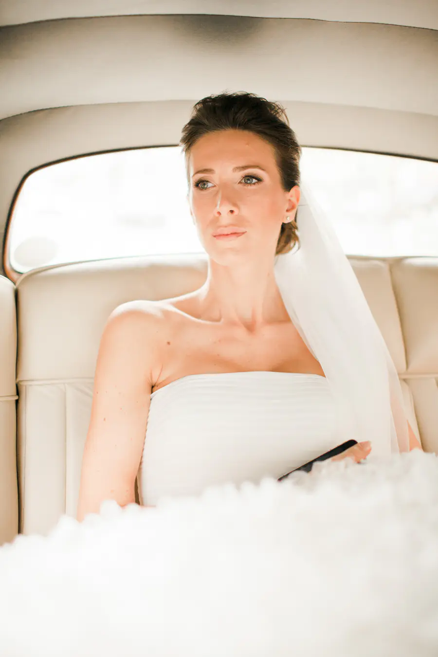 A bride in a white strapless dress and a sheer veil sits in the back seat of a car, looking calmly towards the left. She has brown hair styled up and wears small pearl