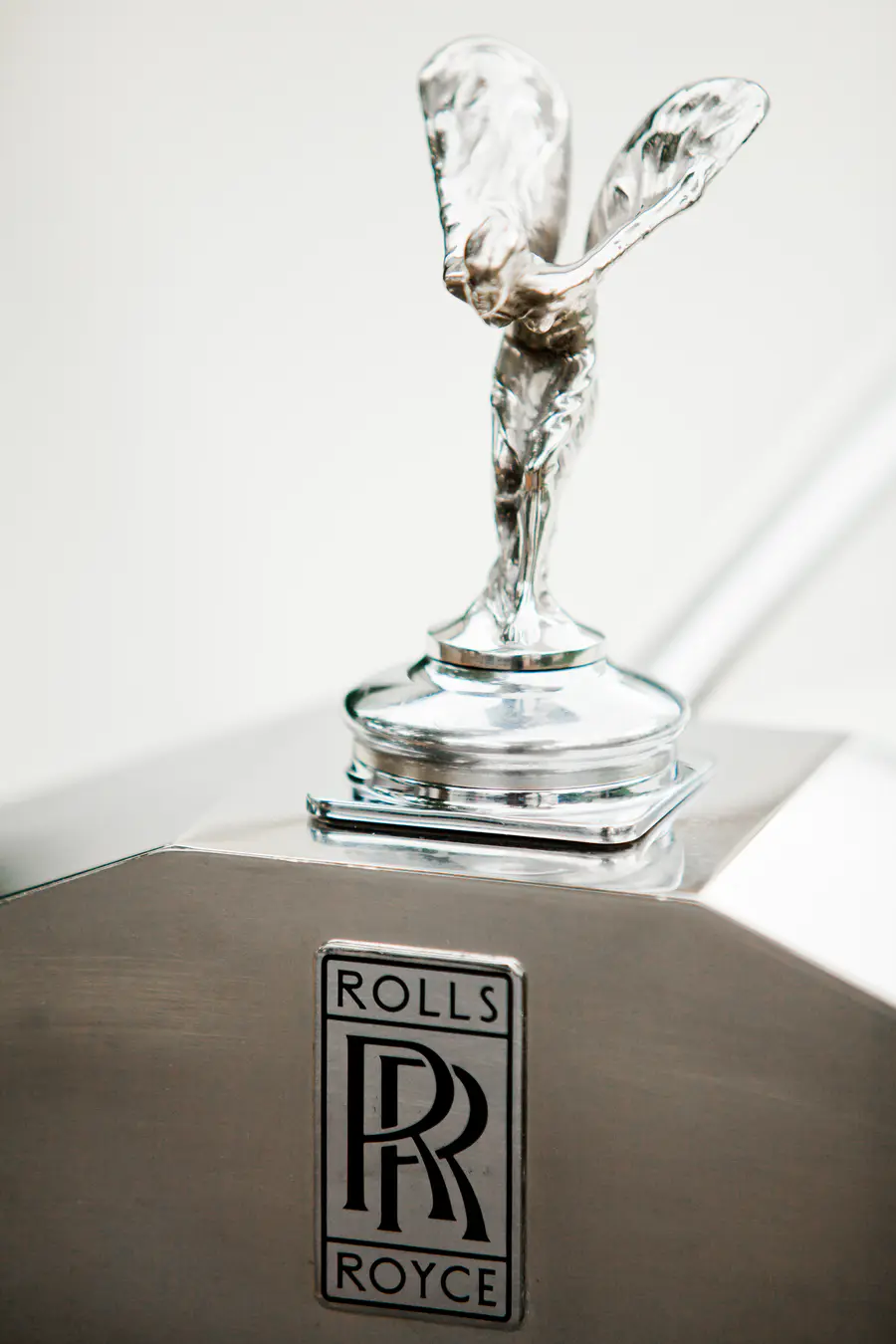A silver Rolls-Royce hood ornament, known as the Spirit of Ecstasy, is pictured in close-up. The emblem is mounted on a polished metal surface, with the "Rolls-Royce" logo visible below it.