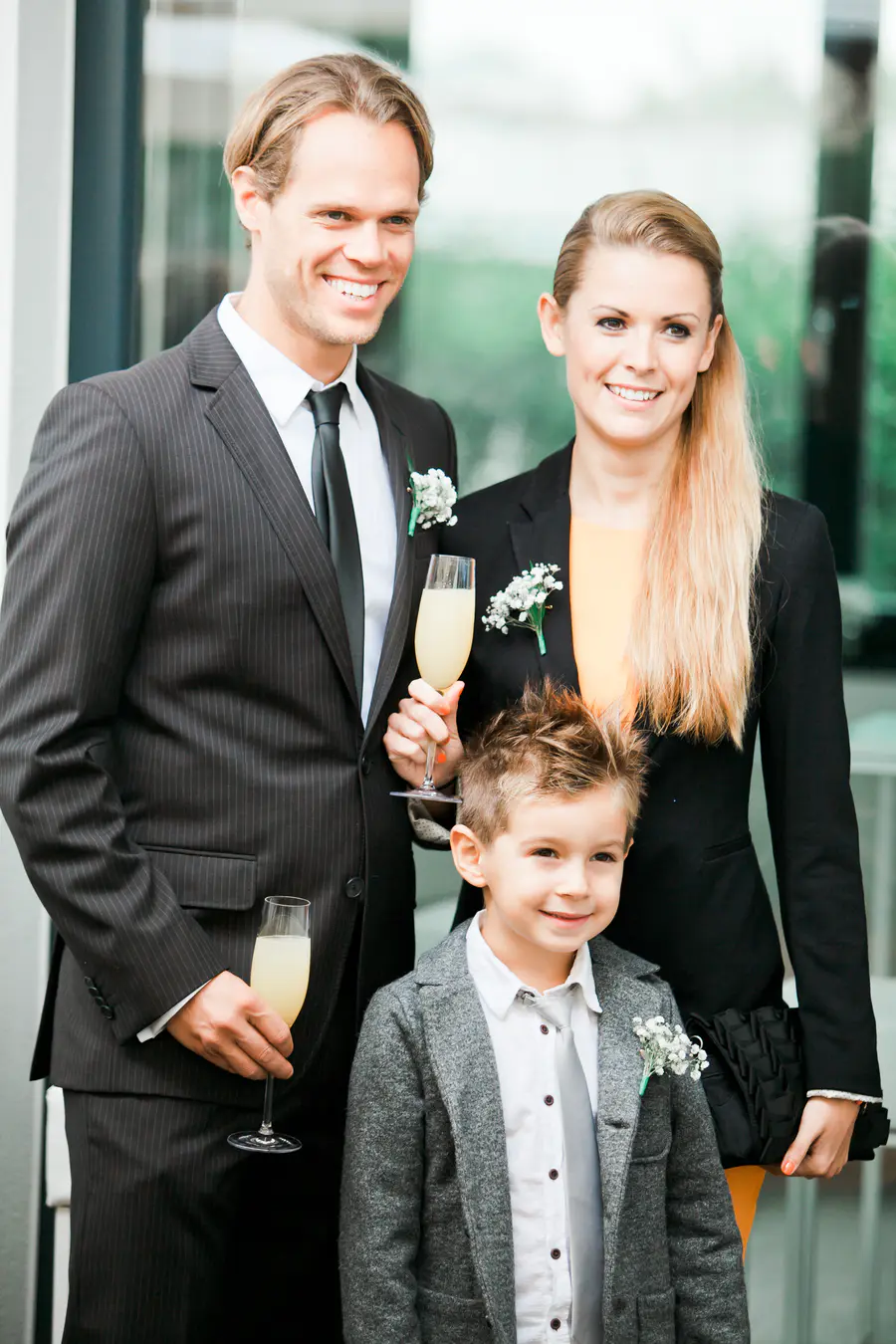 A family of three, two adults and a young boy, pose for a photo. The adults are dressed in formal attire, with the man in a pinstripe suit and the woman in a black suit jacket over an orange top. The boy is wearing a grey blazer over a white shirt and tie. All three are smiling and holding glasses of champagne.