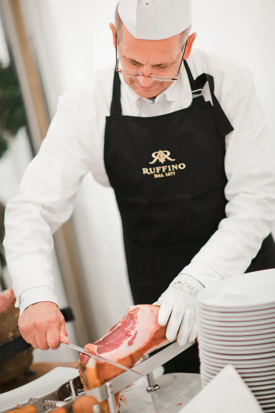 A man wearing a white chef's hat, glasses, and a black Ruffino apron carves a leg of cured ham with a knife on a slicer.