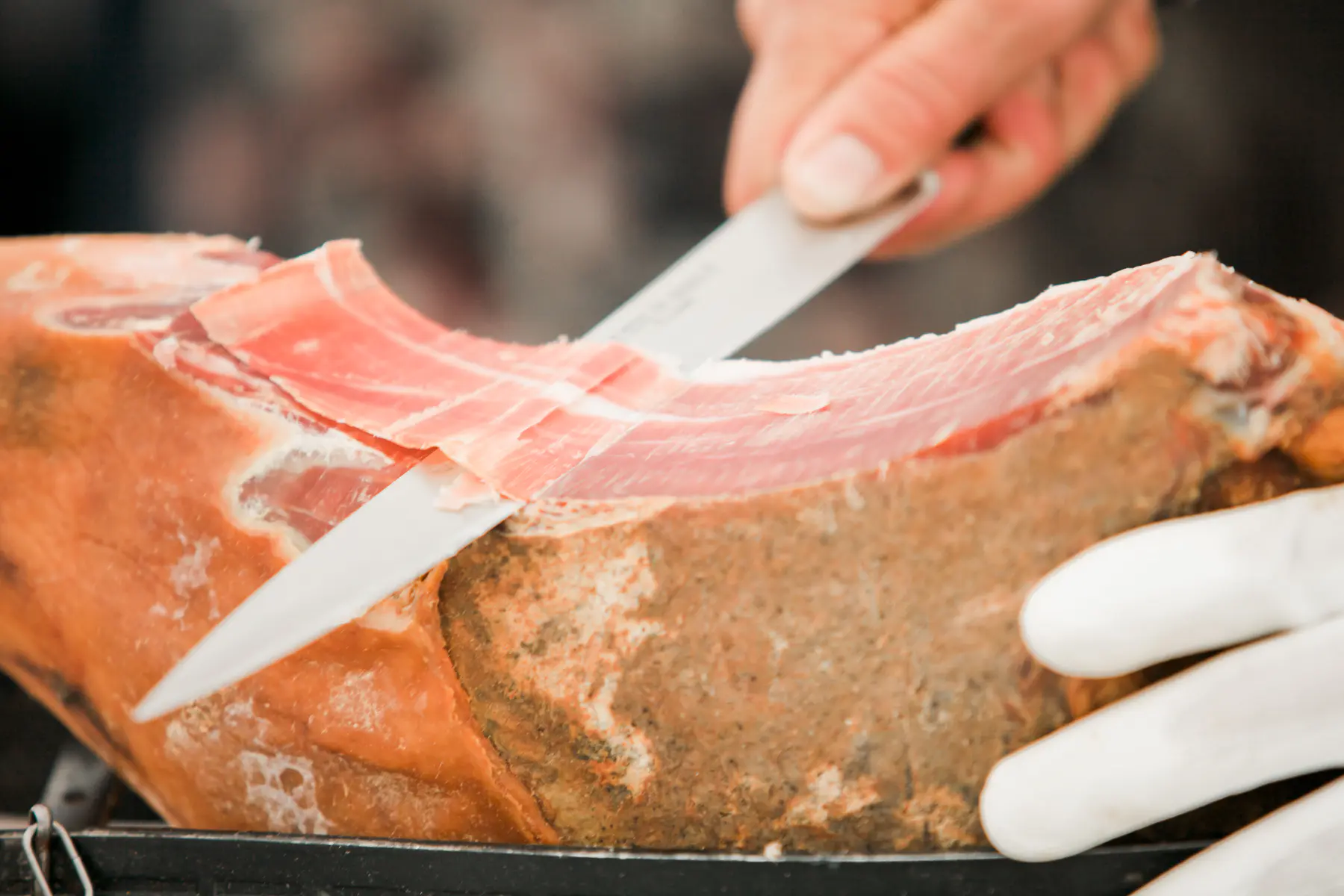 A hand wearing white gloves slices thin strips of cured ham from a large leg of prosciutto with a silver knife.