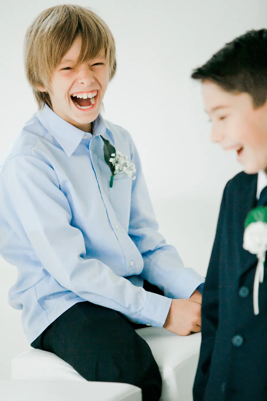 A young boy in a light blue dress shirt laughs heartily, while another boy in a dark suit smiles to his right.
