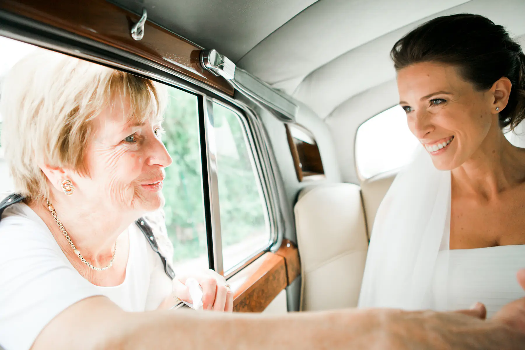 A bride in a white dress smiles at an older woman in a car.