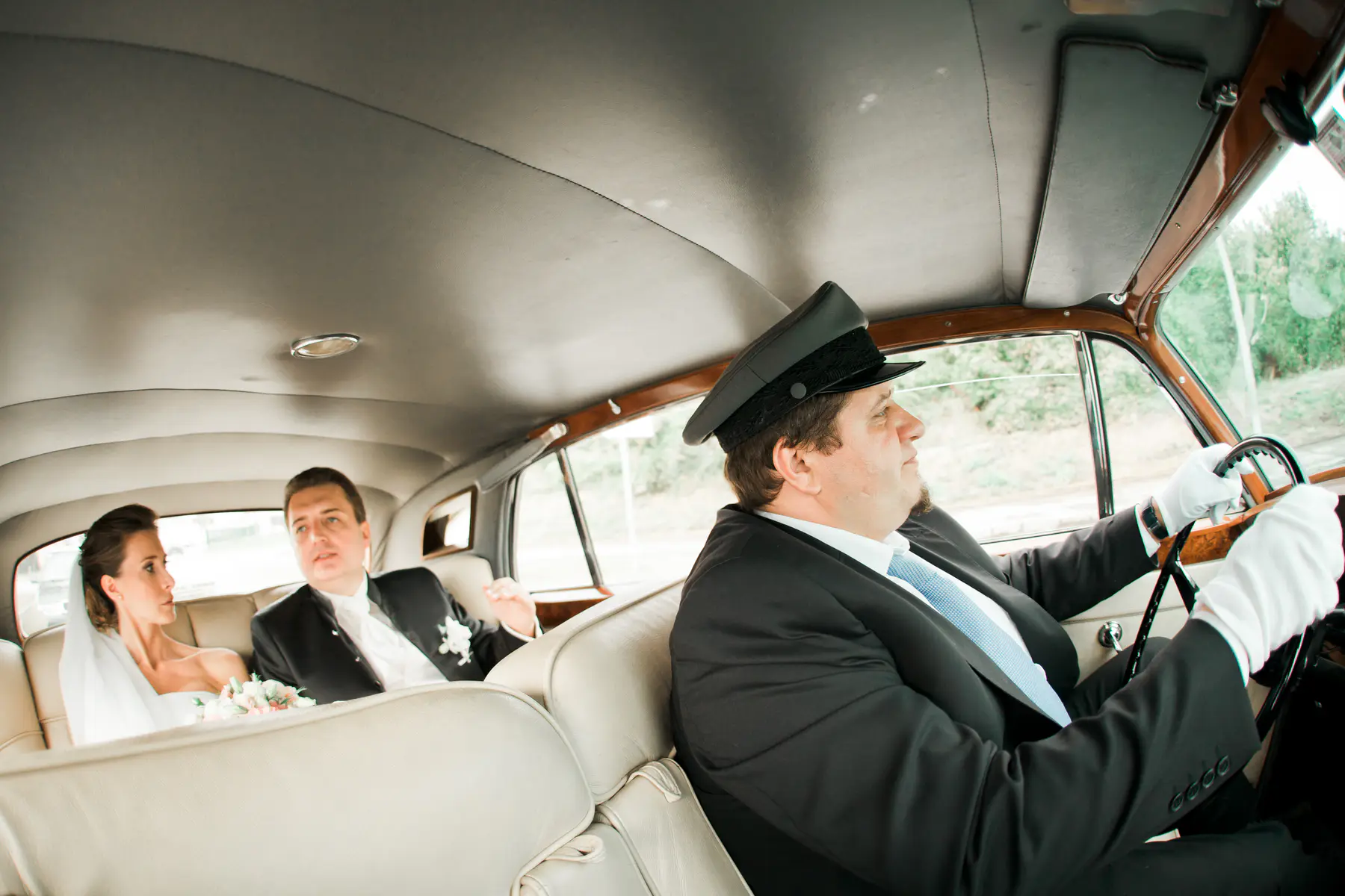 A chauffeur in a hat and white gloves drives a classic car with a bride and groom in the back seat.