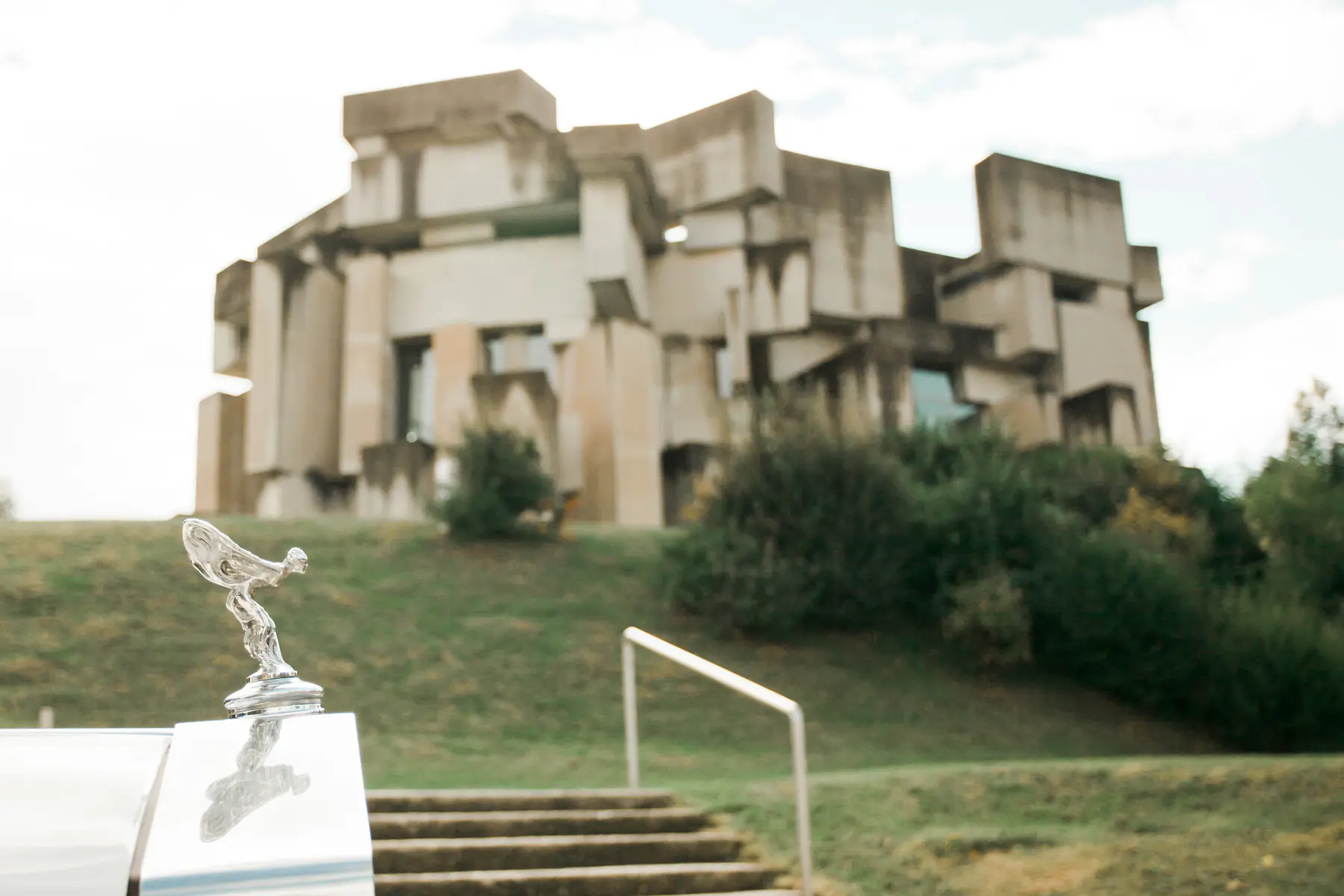 A silver Rolls-Royce hood ornament, the Spirit of Ecstasy, stands prominently in the foreground, its polished surface reflecting the light. Behind it, a set of concrete stairs leads up a grassy slope towards a Brutalist-style building characterized by its angular, block-like concrete structure. Lush green bushes surround the building, and the sky above is a pale, overcast white.