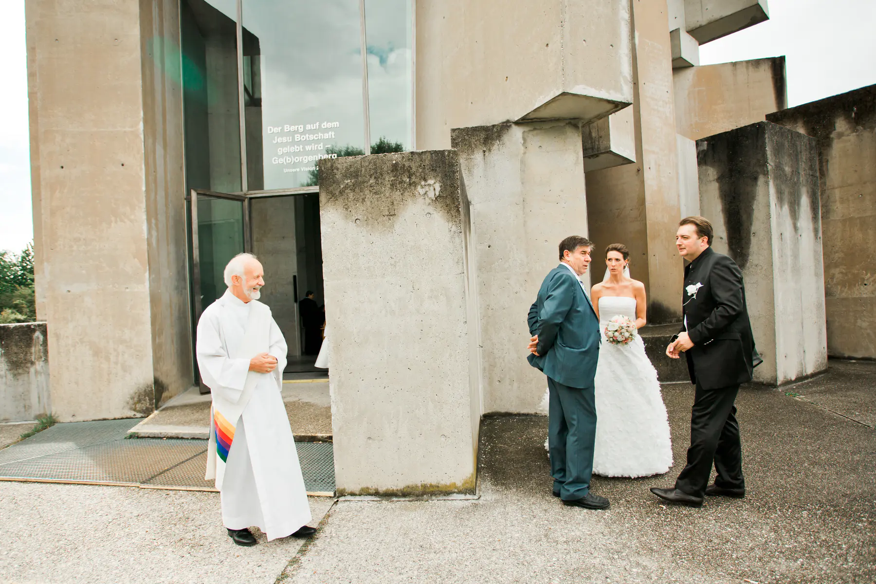 A wedding party gathers outside a modern concrete building. On the left, a priest in white robes smiles, a rainbow sash visible. To the right, the bride in a white strapless gown holds a bouquet, standing between two men in suits.