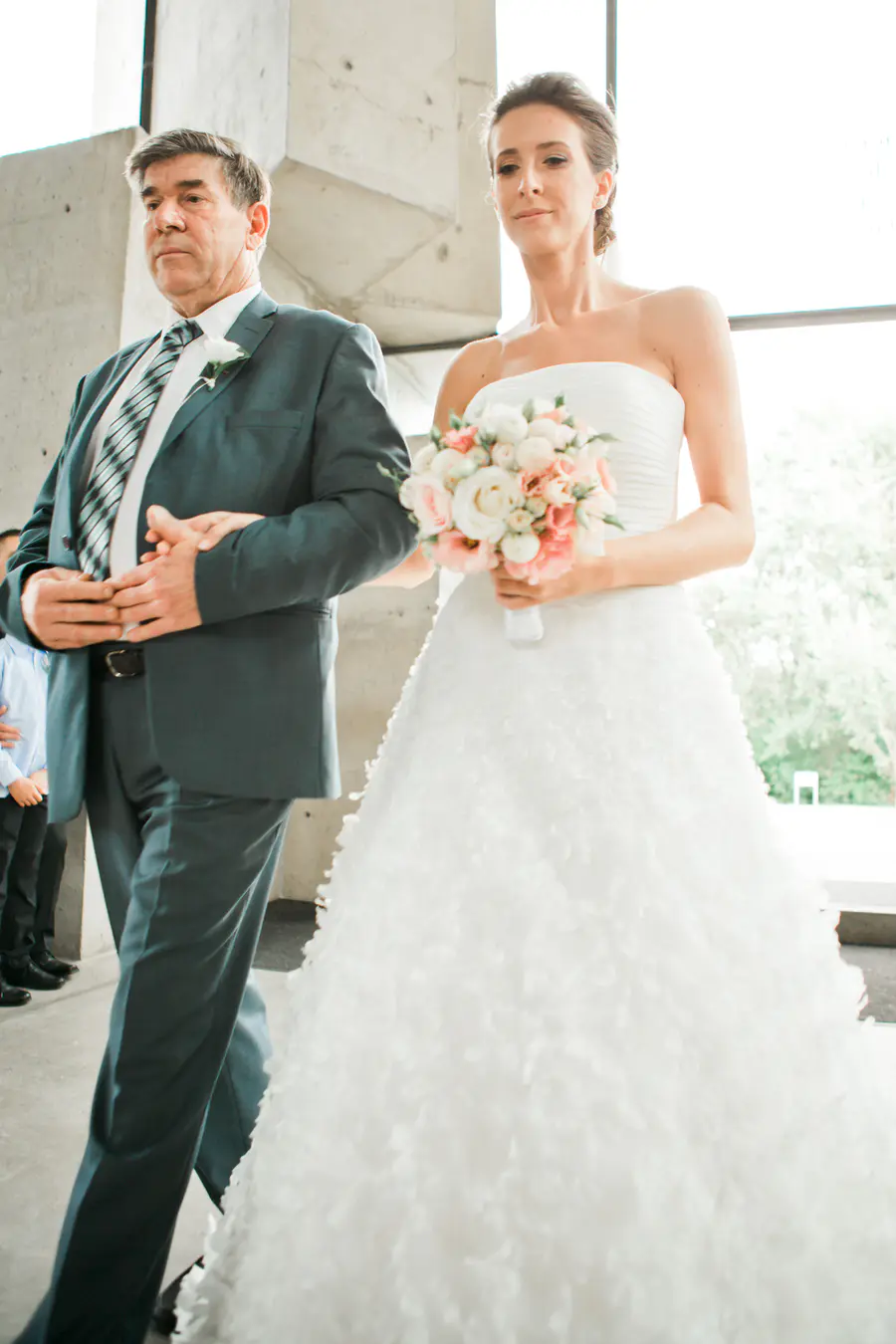 A bride in a strapless white gown walks down an aisle, holding a bouquet of pink and white flowers, while being escorted by a man in a suit.