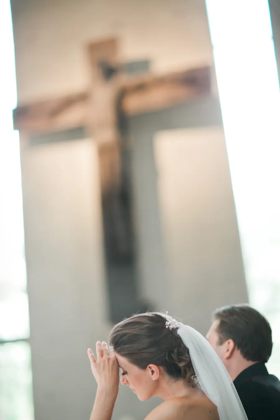 A bride in a white veil bows her head with her hands pressed to her forehead in prayer, in front of a blurred wooden crucifix.