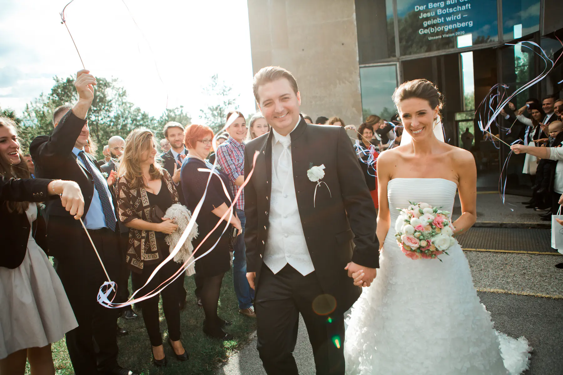 A smiling bride and groom walk hand-in-hand with guests cheering and throwing ribbons.