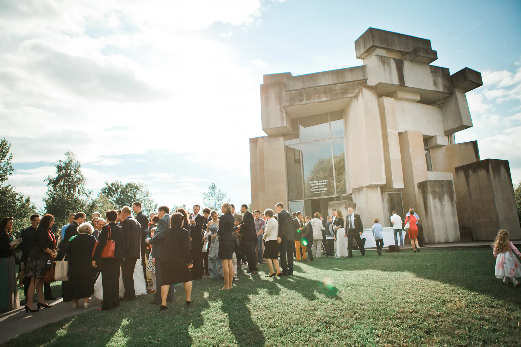 A crowd of people in formal attire gather on a grassy lawn in front of a brutalist concrete building with large windows.