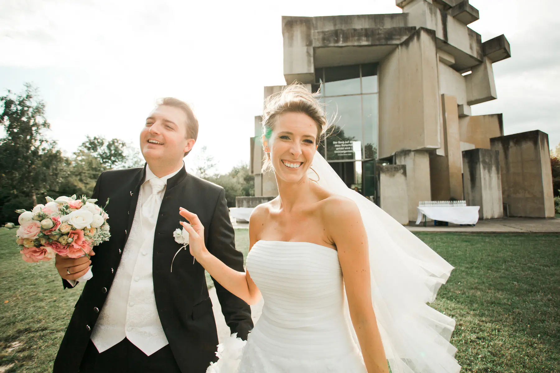 A bride and groom are laughing as they walk away from the camera in front of a Wotruba Church (Kirche Zur Heiligsten Dreifaltigkeit)