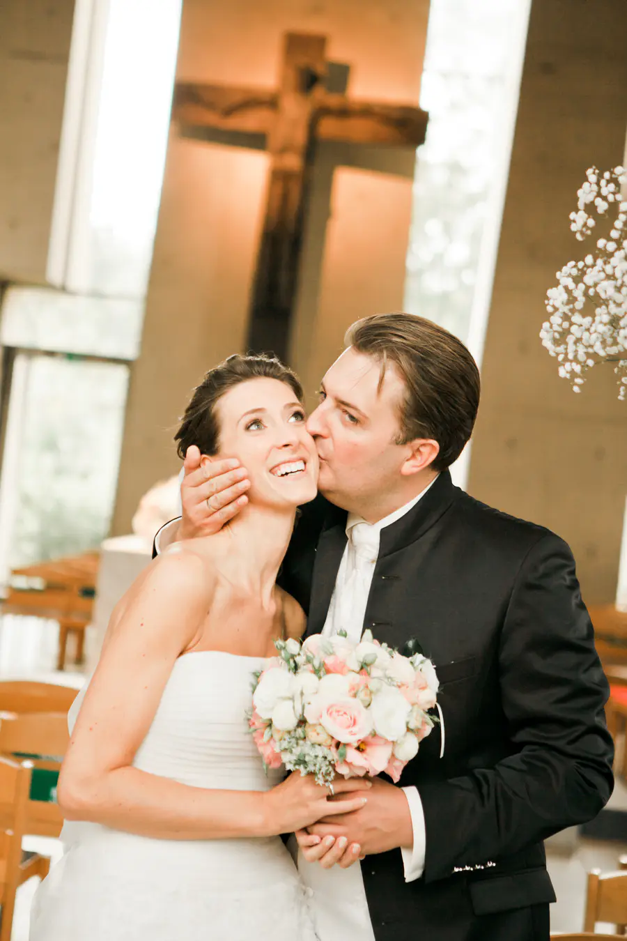 A groom kisses his bride on the cheek while she smiles and looks up, holding a bouquet of pink and white roses.