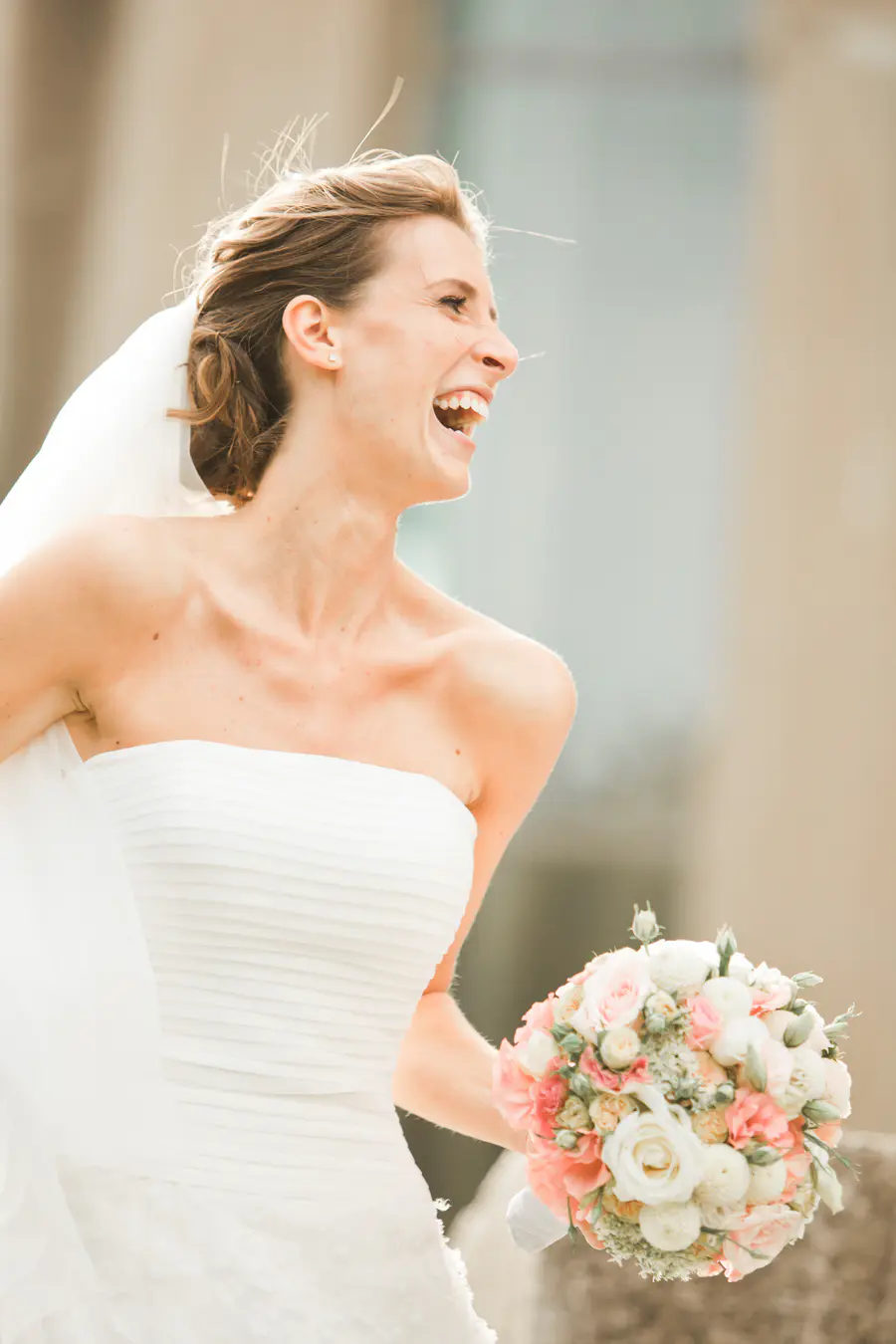 A bride in a strapless white wedding dress laughs joyfully while holding a bouquet of pink and white roses and greenery.