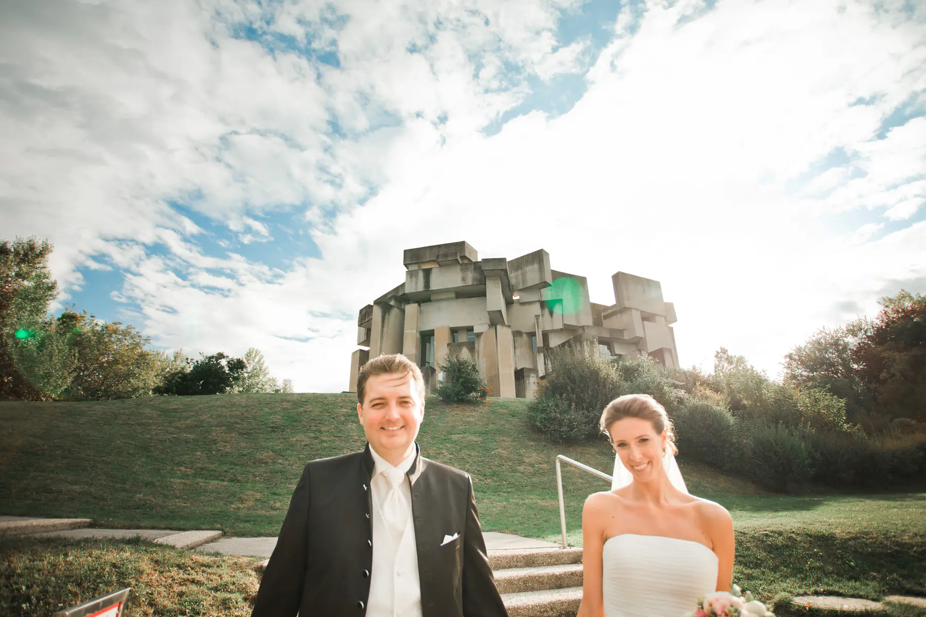 Capturing timeless silhouettes at the Wotruba Church. A smiling bride and groom stand on concrete steps outdoors, with a unique angular brutalist concrete building on a grassy hill behind them under a bright, partly cloudy sky.
