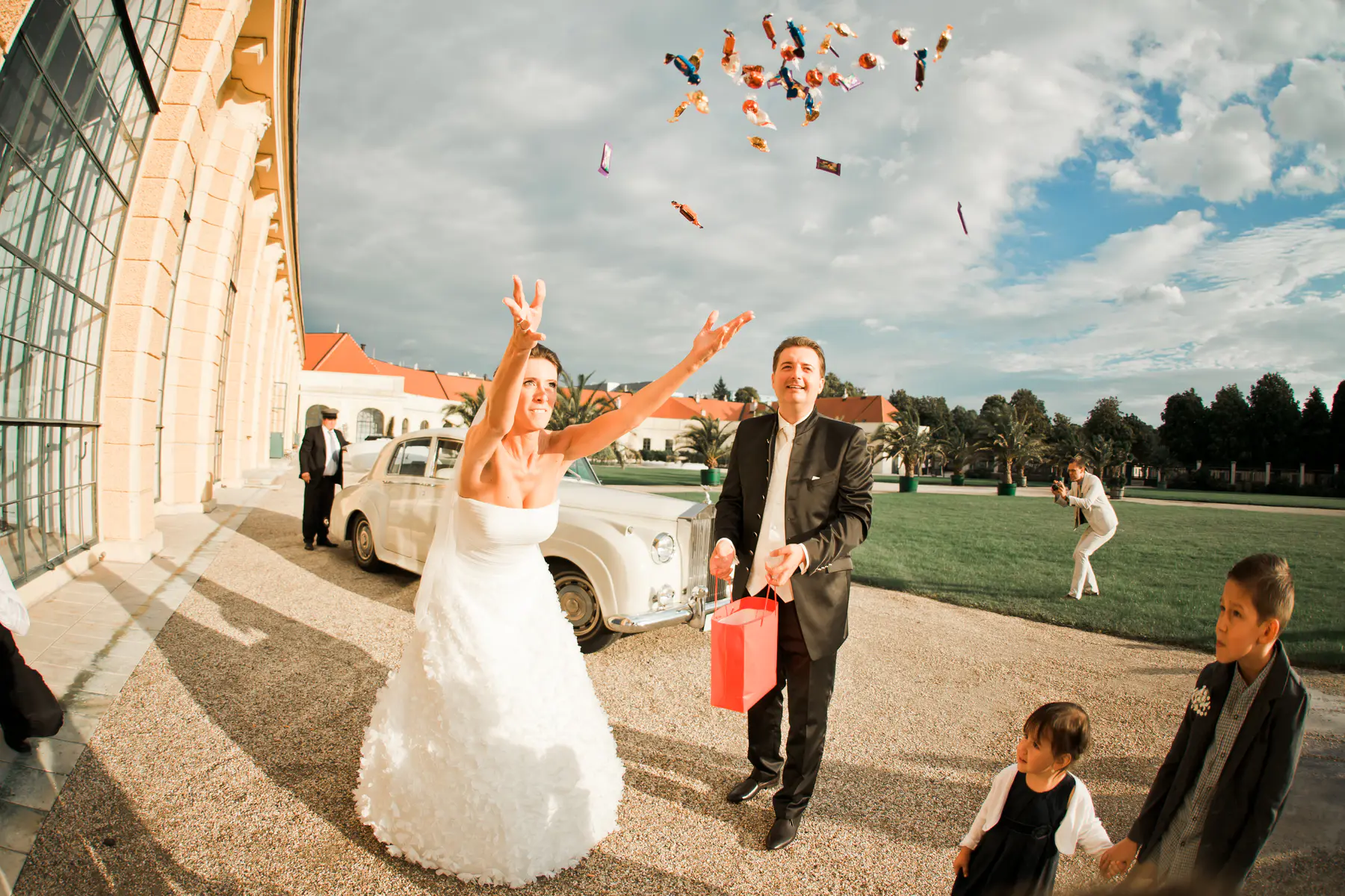 A smiling bride in a white dress and a groom in a black suit throw colorful wrapped candies.They are outside a grand,