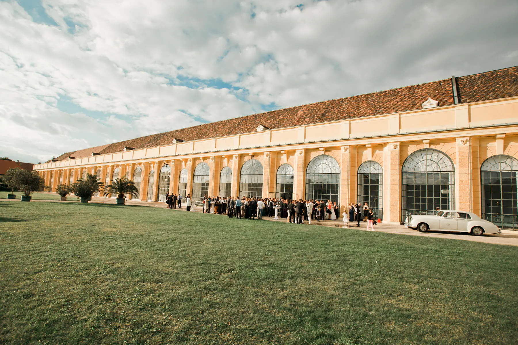 The Orangery at Schönbrunn Palace.A large group of formally dressed people are gathered on a green lawn in front of a long, elegant pale yellow building with many arched windows. A white vintage car is parked to the right. The sky above is