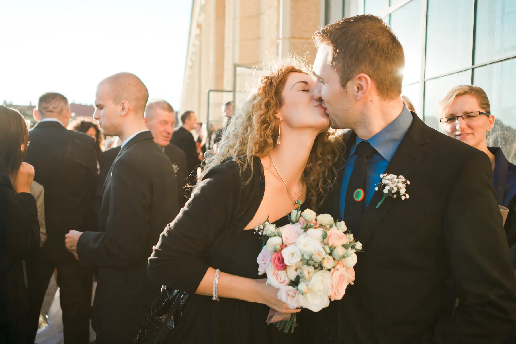 A formally dressed couple shares a tender kiss, bathed in bright sunlight. The woman, with long curly hair, holds a bouquet of light pink and white roses as she kisses the man on the cheek.