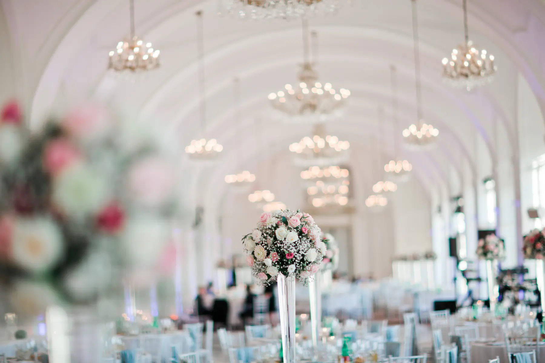The Orangery at Schönbrunn Palace. An elegant event reception hall is decorated with a tall centerpiece of pink and white flowers and baby's breath in the foreground. The long, white vaulted ceiling is lined with many sparkling chandeliers, and tables with light blue chairs are set up throughout