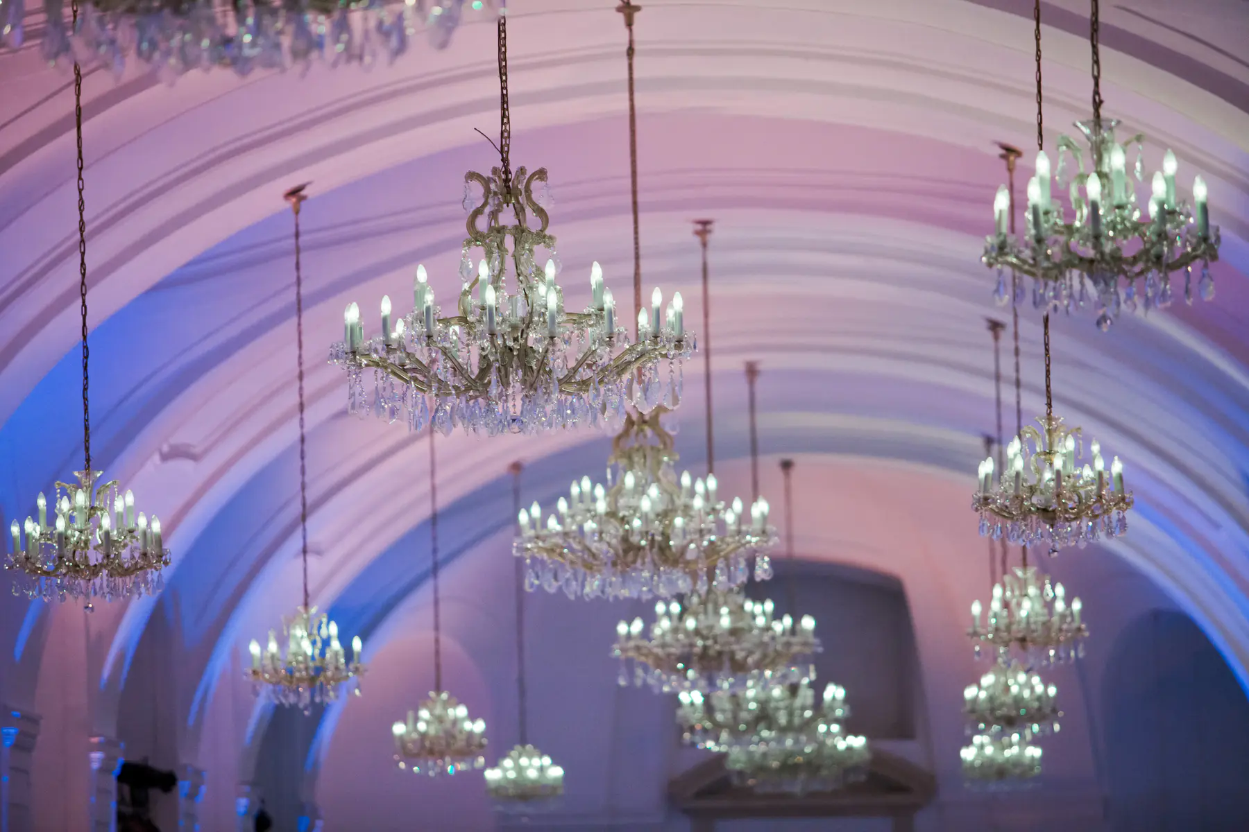 The Orangery at Schönbrunn Palace.Numerous ornate crystal chandeliers with glowing candle-like bulbs hang from a high vaulted ceiling bathed in a gradient of soft purple and blue light.