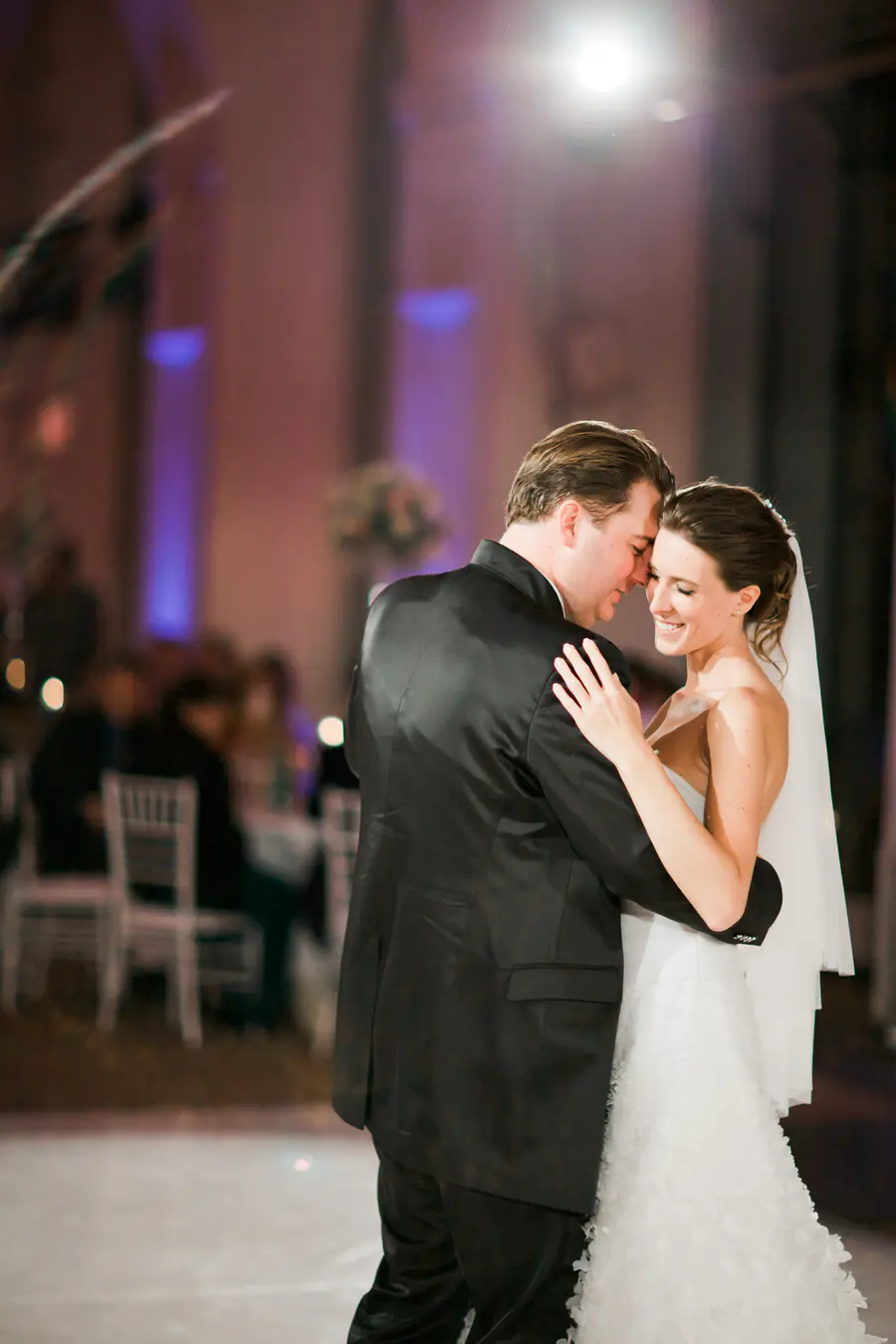 A smiling bride in a white gown and veil dances closely with her groom in a black tuxedo. Their heads are near touching, and the blurred background shows a wedding reception hall with purple uplighting.