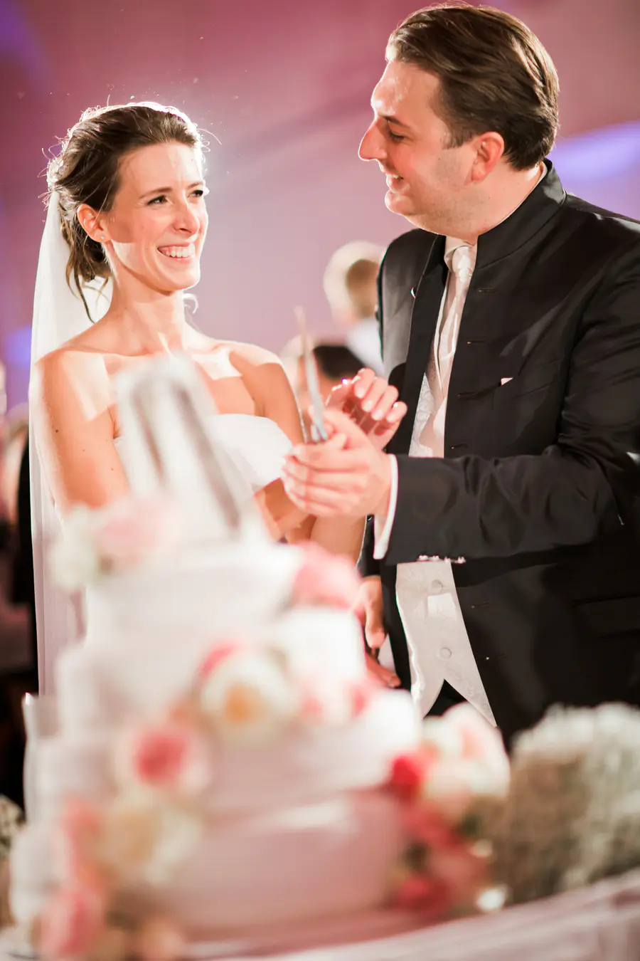 A bride and groom smiling at each other while cutting a multi-tiered wedding cake decorated with pink flowers.