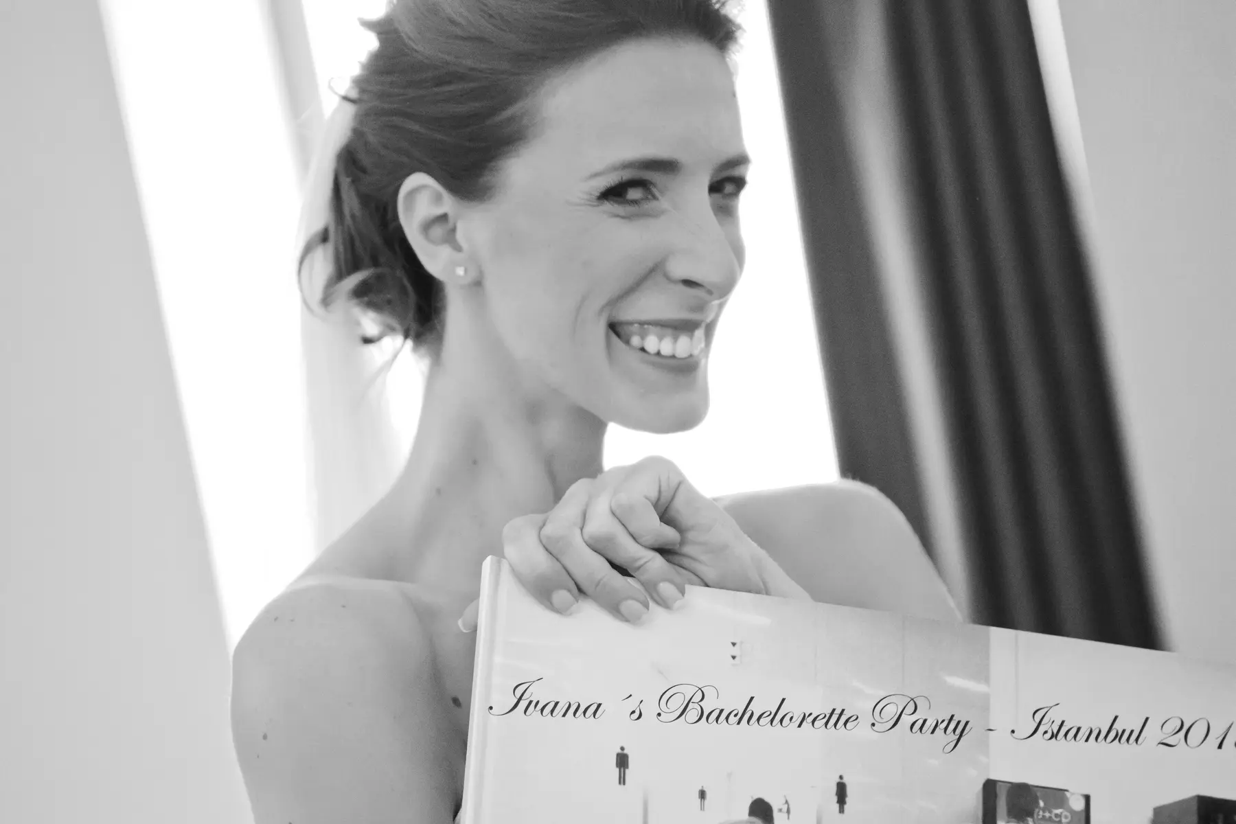 A beaming woman with bare shoulders smiles widely while holding open a book titled "Ivana's Bachelorette Party - Istanbul " in this black and white photo.