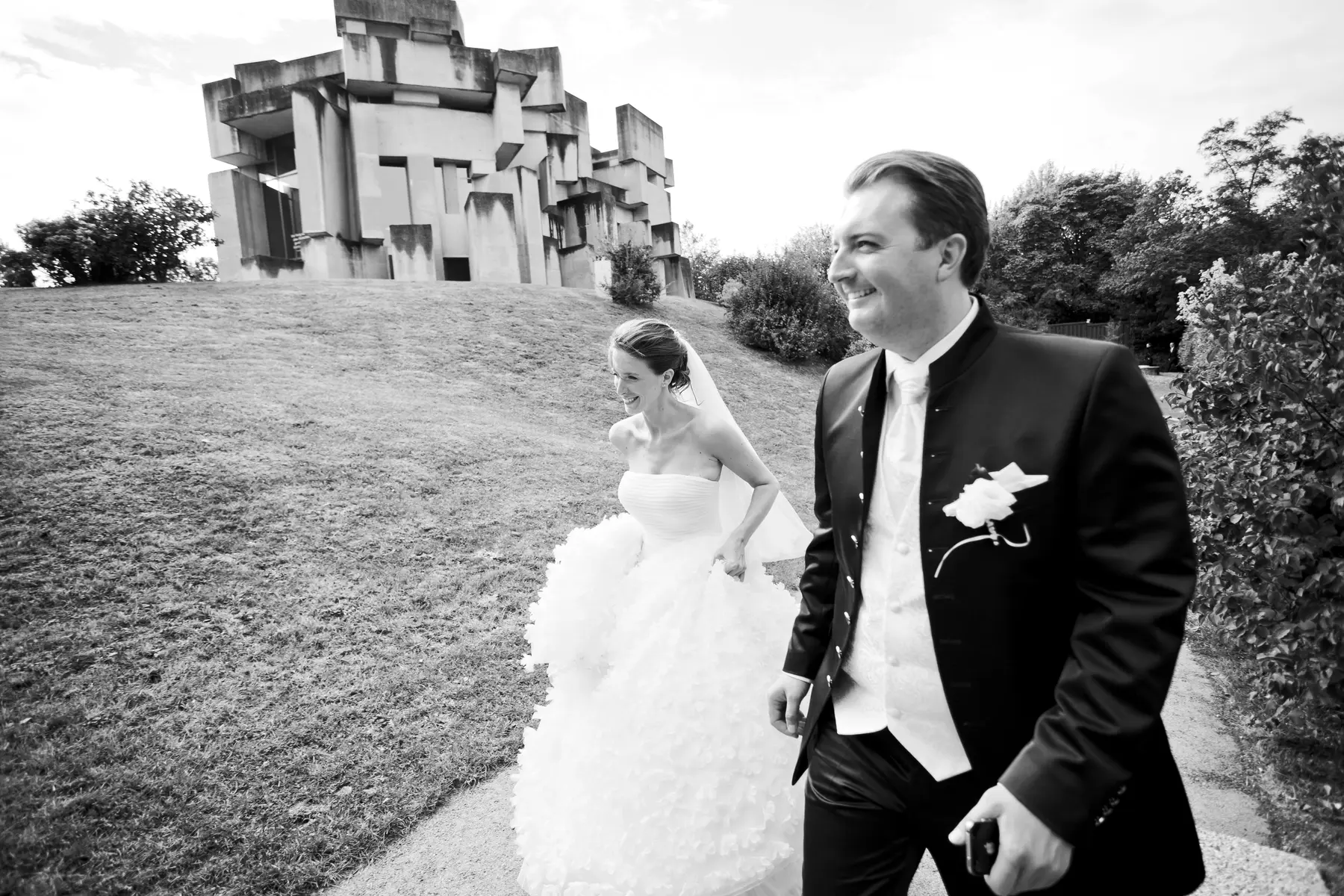 A black and white photo shows a newly married couple walking outdoors. The groom, in the foreground on the right, smiles and looks to his left while holding a small object in his hand. He wears a black suit with a white vest and tie and a boutonniere. Behind him and to the left, the bride walks, looking to her left and smiling. She wears a strapless white wedding gown with a full skirt and a veil. Behind them, a grassy hill slopes up towards a large, angular, concrete structure.