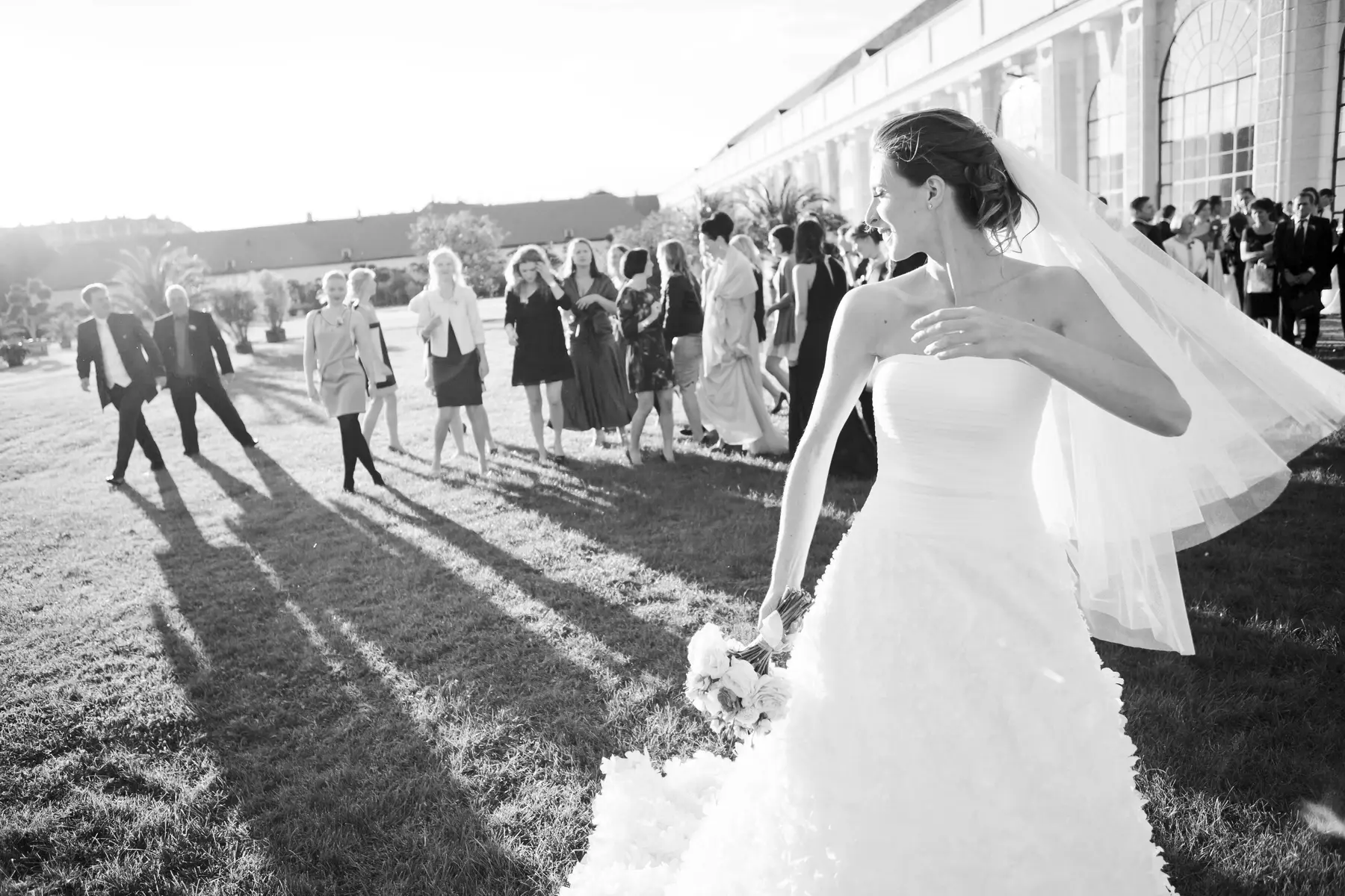 Black and white photo of a smiling bride in a strapless white gown and flowing veil, holding a bouquet. She looks back over her right shoulder at a group of guests gathered on a sunny grassy