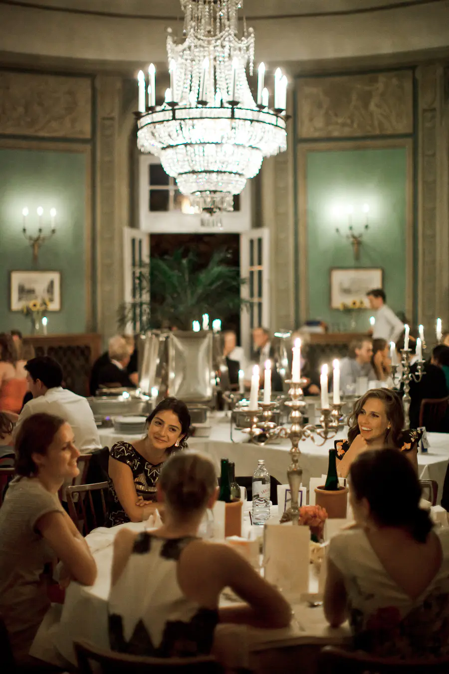A group of people are seated around a table in a dimly lit room, under a large, ornate chandelier.