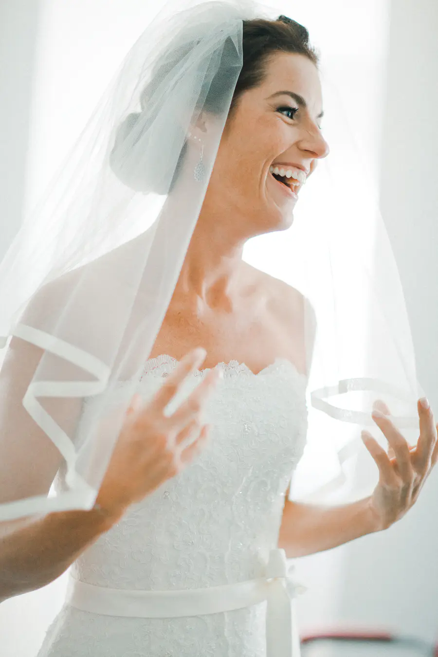 A joyful bride in a white lace wedding dress laughs while holding her sheer veil.