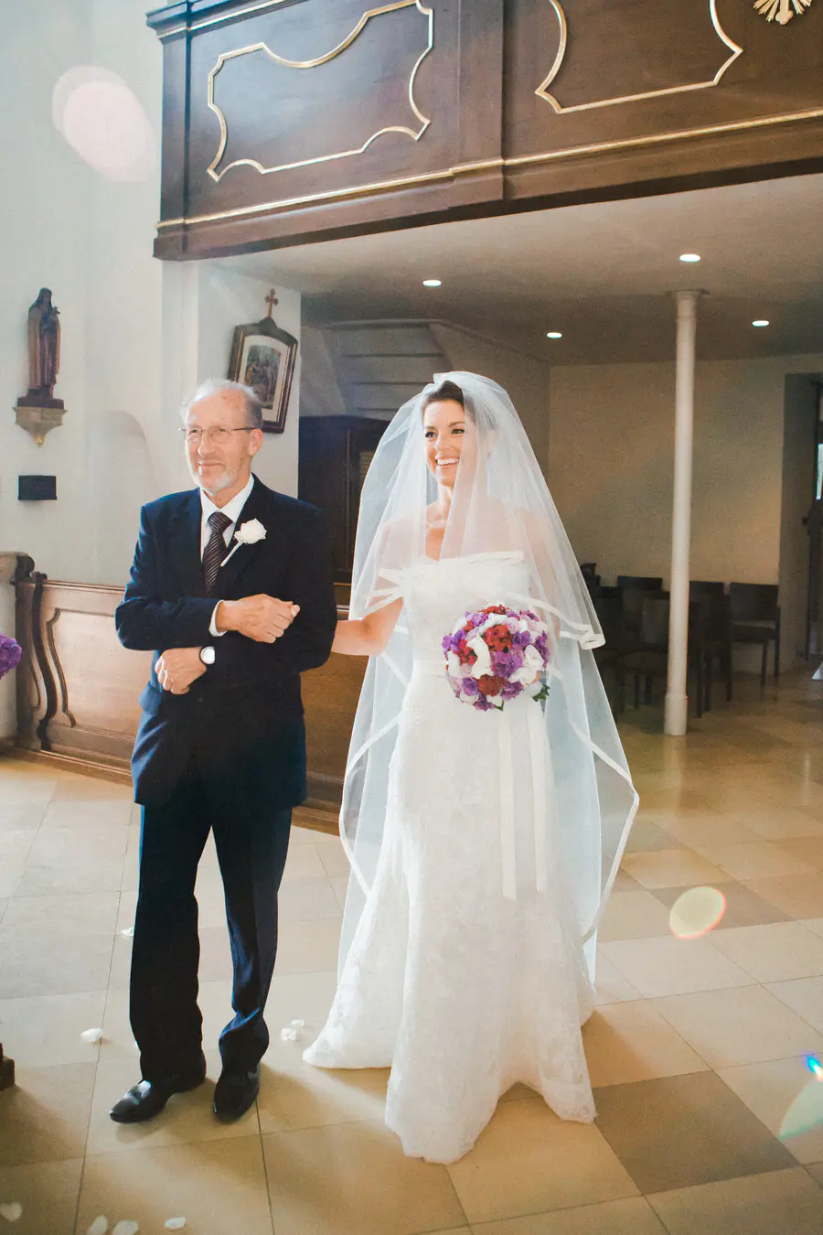 A radiantly smiling bride in a white strapless lace gown and long sheer veil walks arm-in-arm with an older man in a dark suit down a church aisle. She holds a purple and red wedding bouquet. White petals are scattered on the tiled floor, and an ornate wooden balcony is visible in the background.