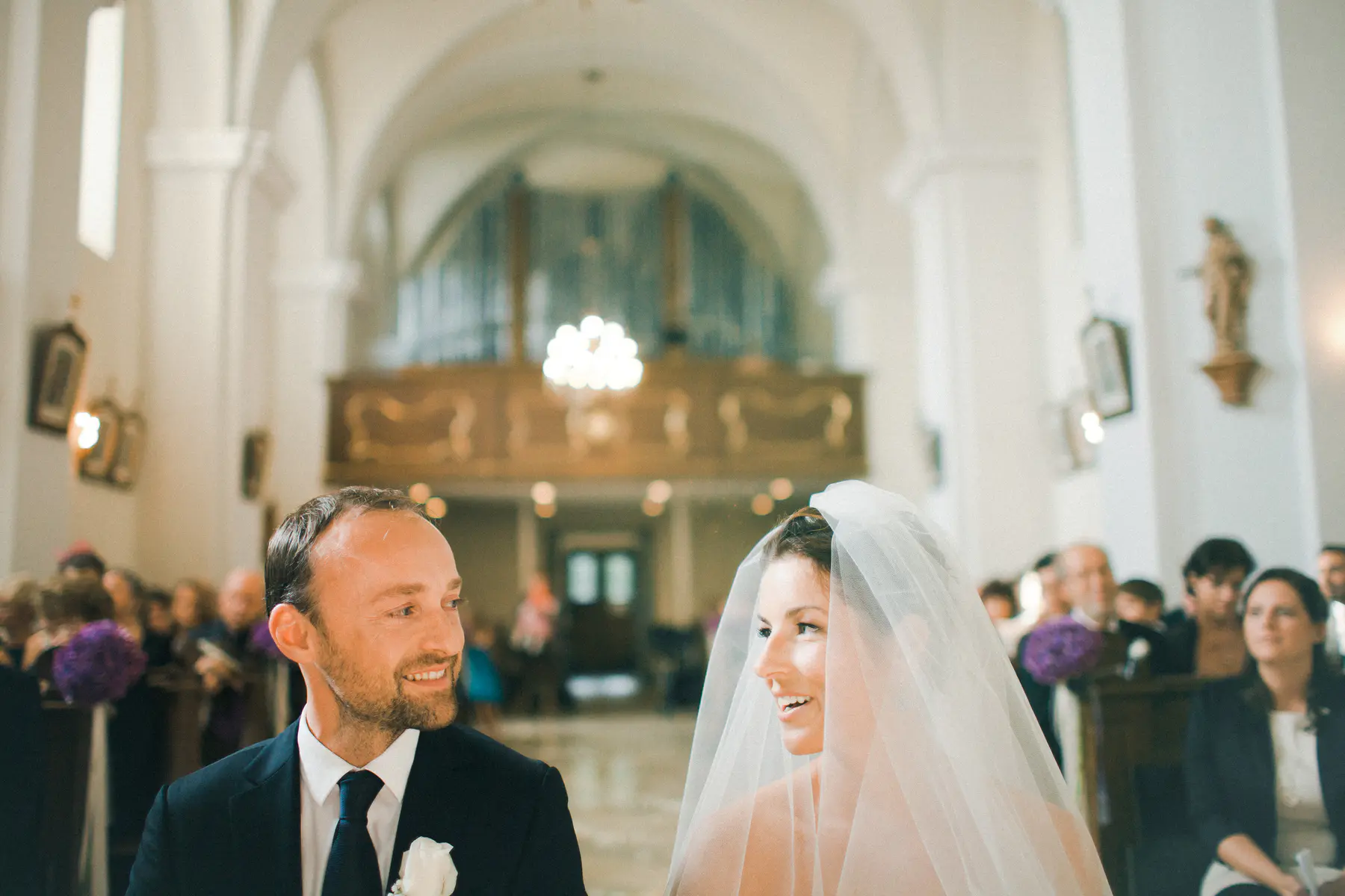 A smiling bride and groom, both in their 30s, gaze at each other during their wedding ceremony inside a church at Schloss Halbturn with high arched ceilings and an organ loft in the background. The groom wears a dark suit and a white boutonnière, and the bride wears a white veil.